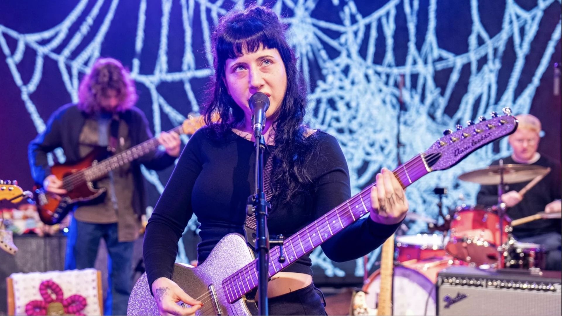 Female musician with black hair and black outfit playing a sparkly purple guitar and singing into a microphone, with a bassist and drummer behind her and a spiderweb backdrop.