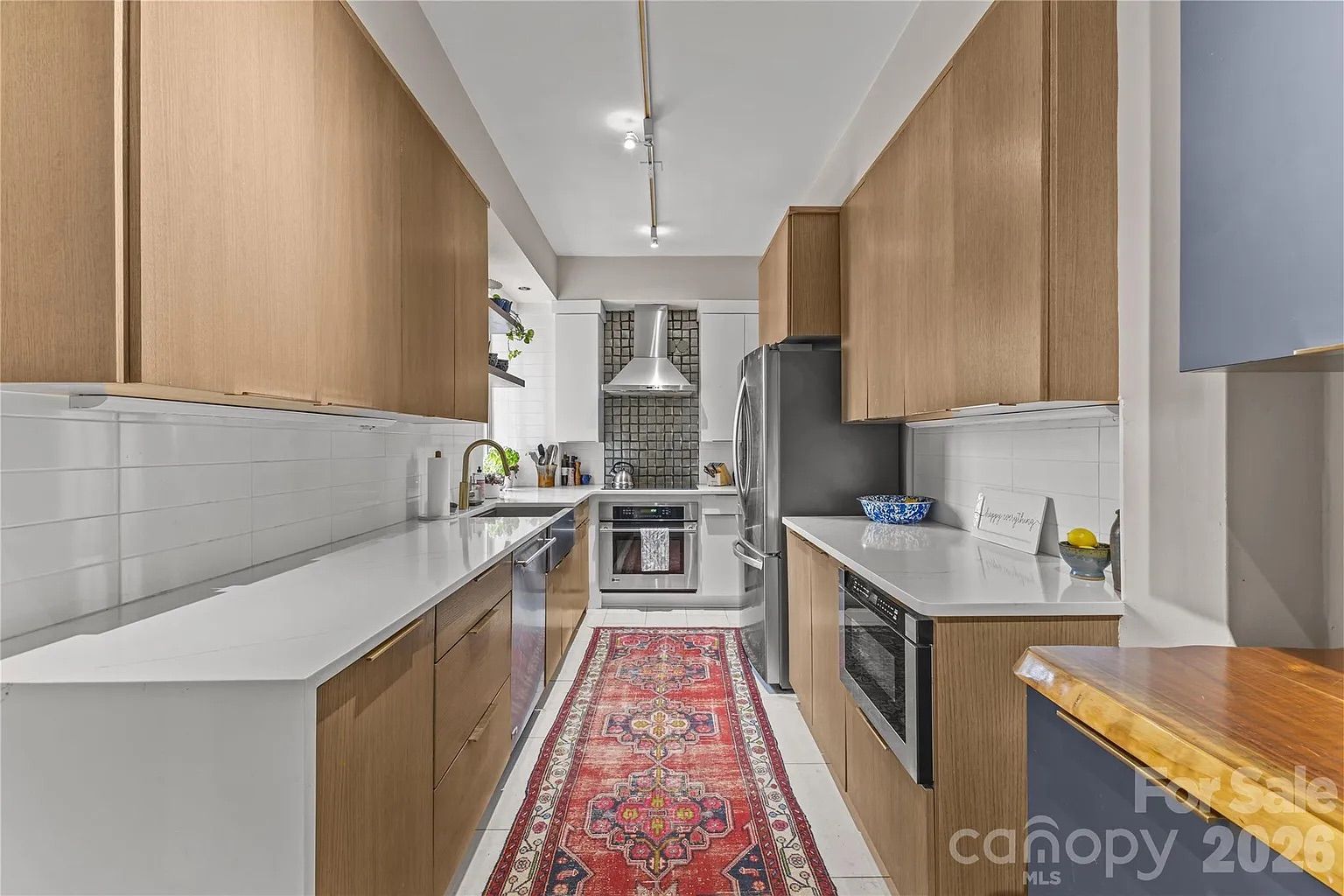 Narrow modern kitchen with wood cabinets, white countertops, stainless steel appliances, a red patterned runner rug, and white tiled backsplash with a dark mosaic behind stove.