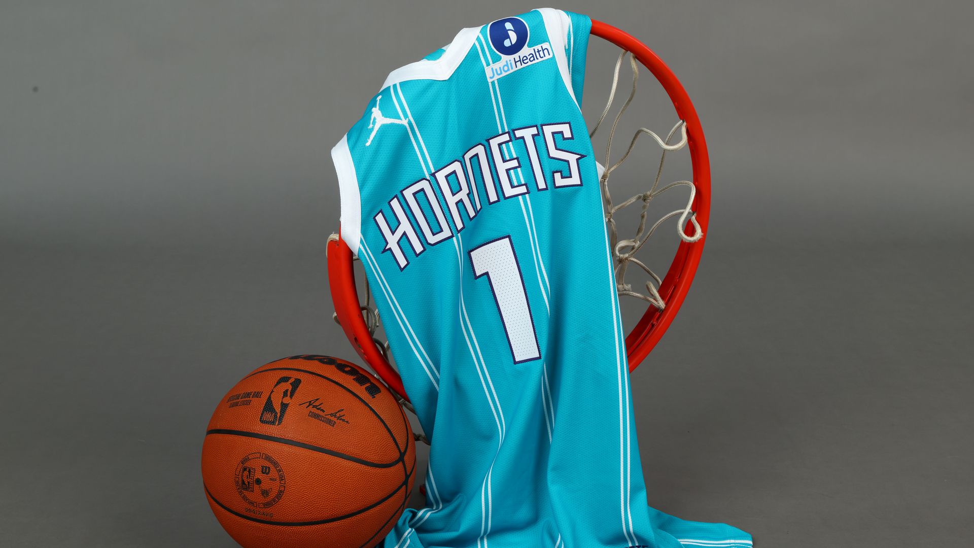 Teal Charlotte Hornets basketball jersey with number 1 draped over an orange basketball hoop rim, next to an official NBA basketball on a gray background.