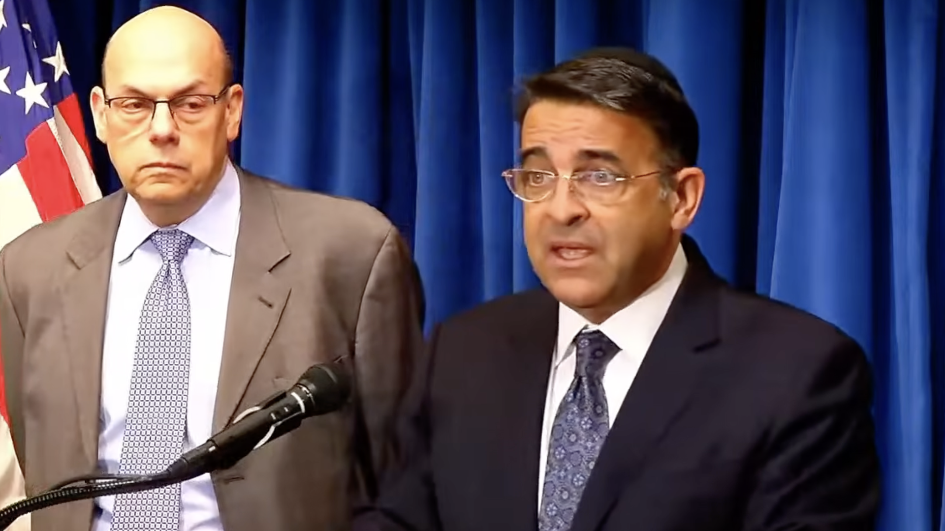 Two suited men at a press conference; left is bald with glasses, right has dark hair and glasses. US flag and blue curtains behind them.