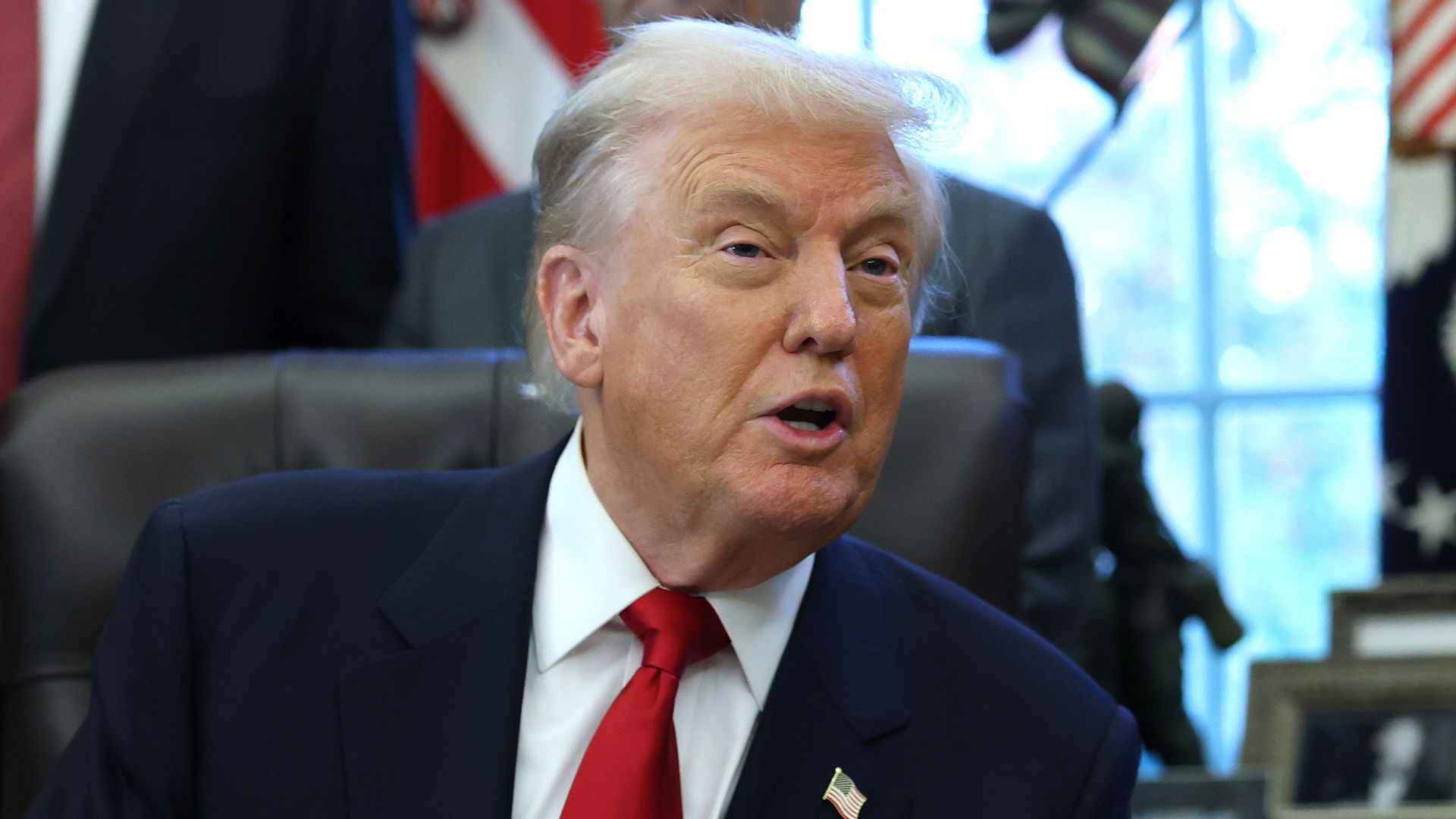 President Trump, wearing a navy jacket with a U.S. flag pin on it, white shirt and red tie, speaks from a black leather chair in the Oval Office of the White House.