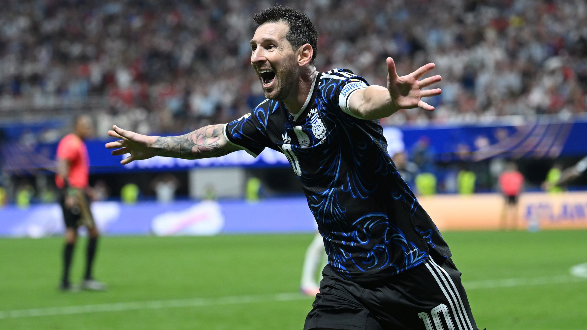 Male soccer player in a blue and black jersey celebrates with arms outstretched on a green pitch, crowd blurred in the stadium behind him; number 10 on his shorts.
