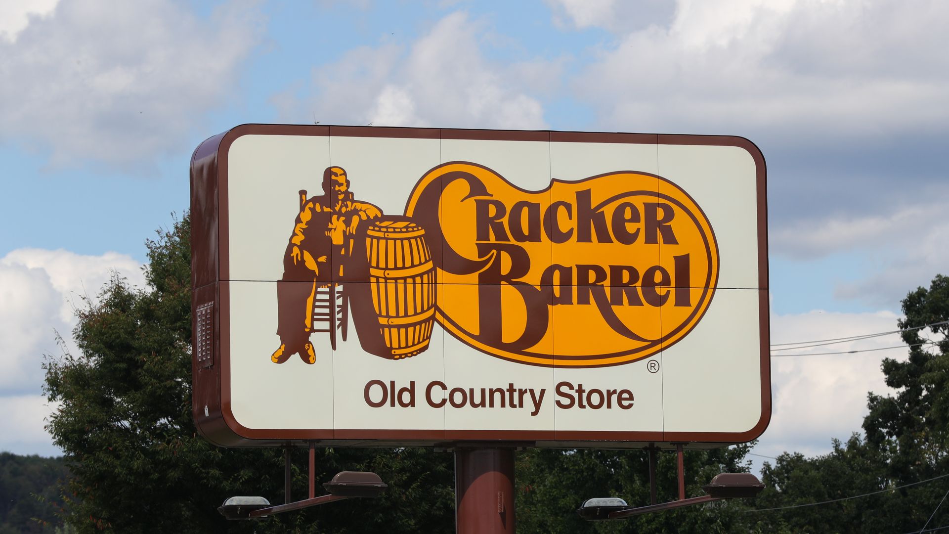 Sign with Cracker Barrel Old Country Store logo featuring a man sitting on a chair beside a barrel, brown and orange colors, trees and sky background.