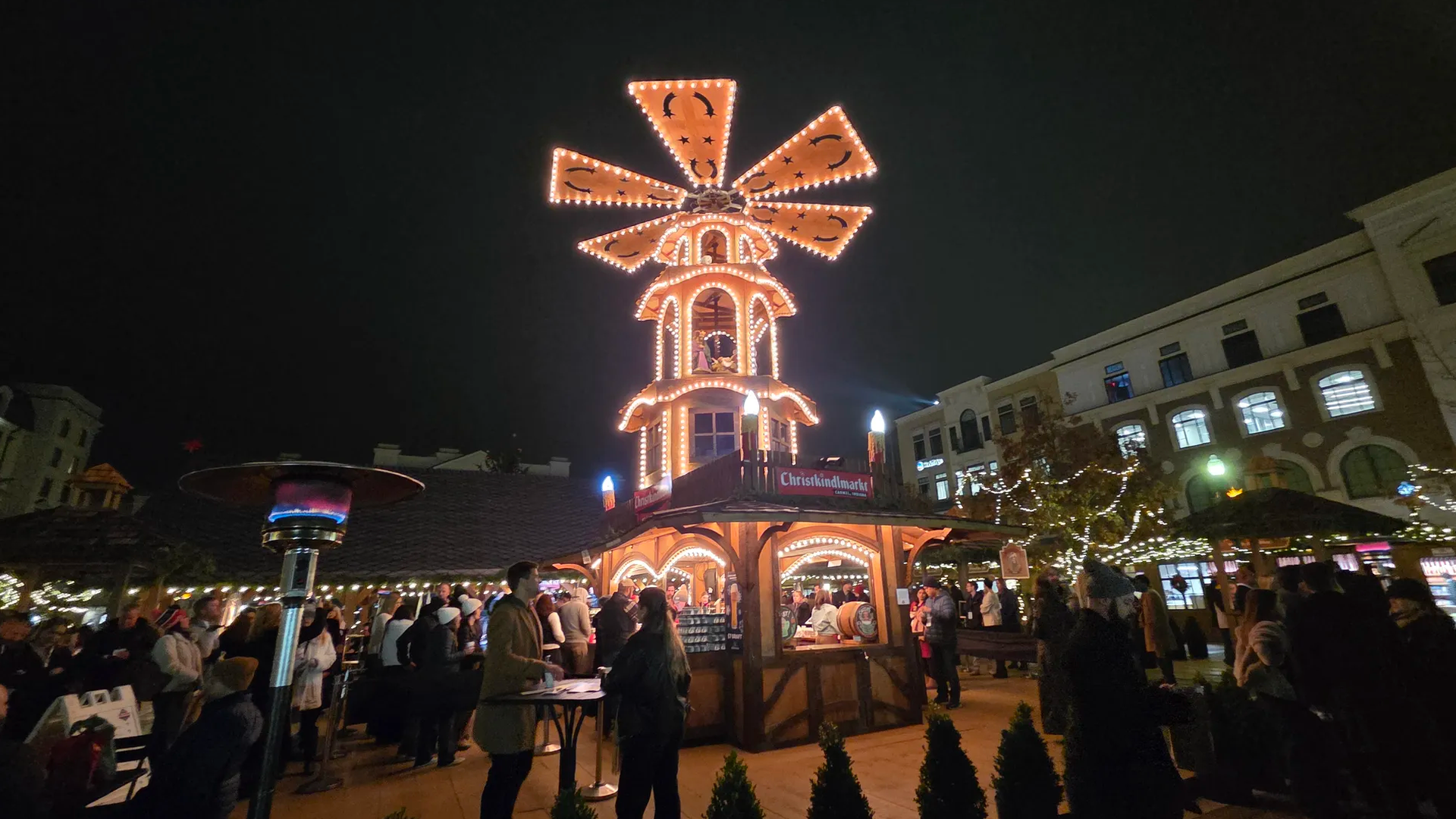 Night scene of a Christmas market with a tall, orange-lit windmill centerpiece atop a decorative pavilion, string lights, people at stalls, and surrounding illuminated buildings.