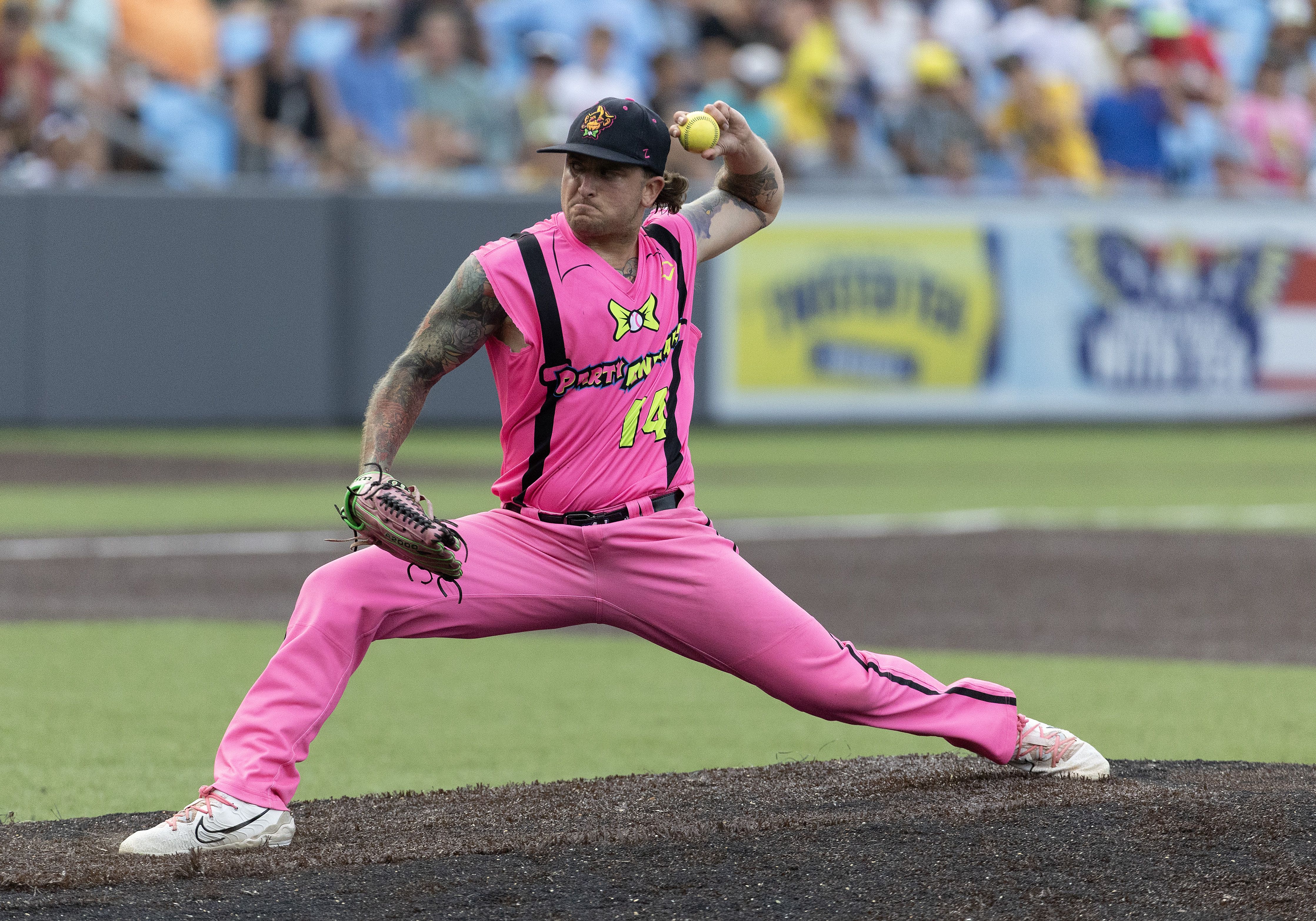 A baseball player for the Party Animal steps toward home plate to pitch a ball with a cut off jersey top and glove in hand.
