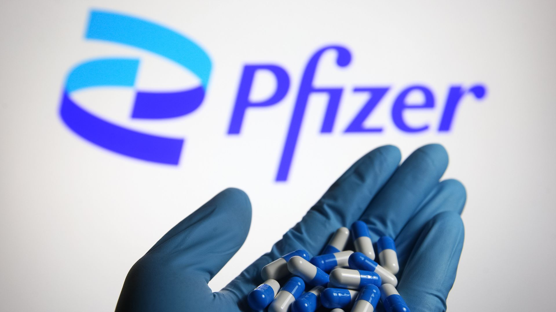 The blue Pfizer logo in the background, and a glove holding blue and white pills in the foreground.