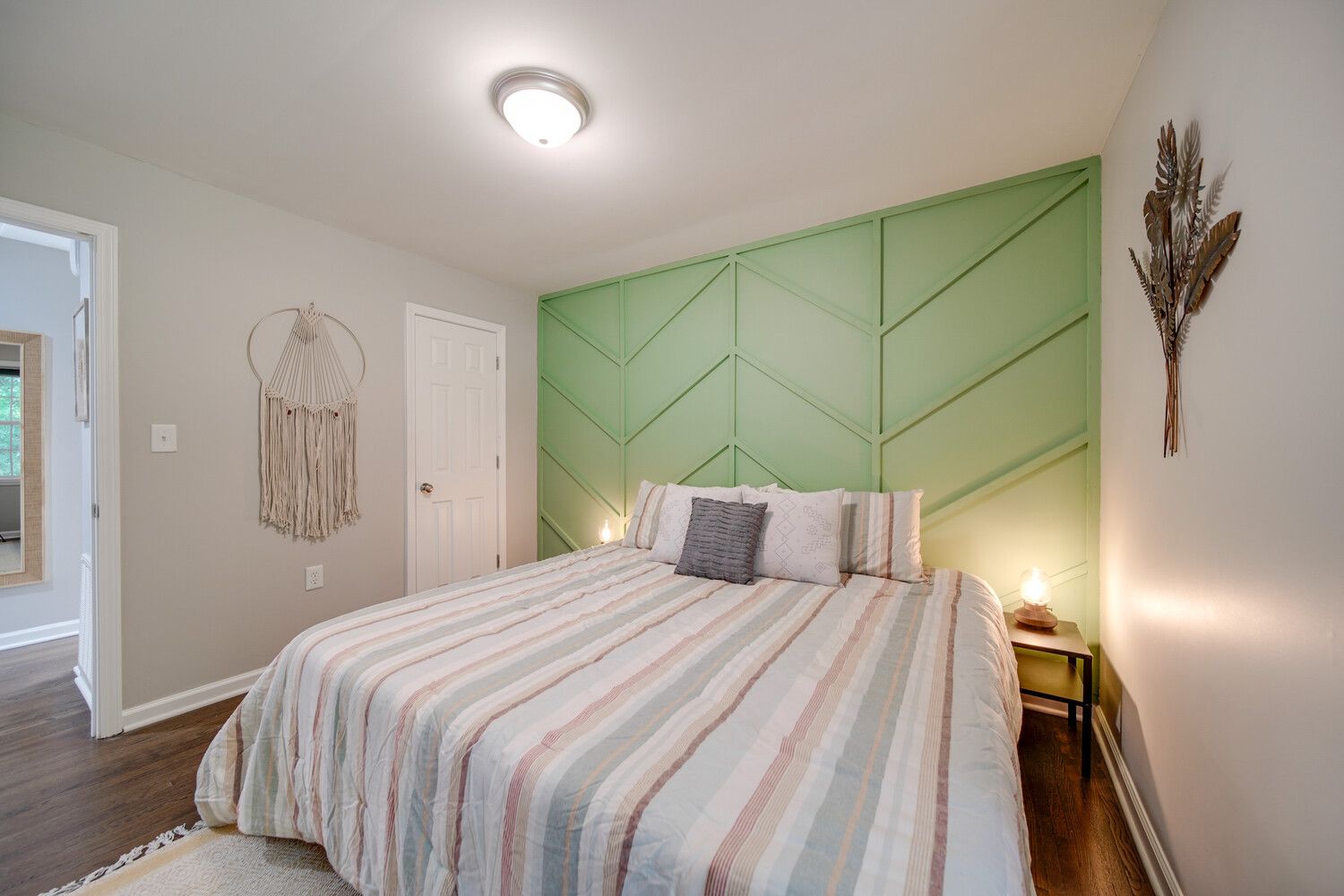 Bedroom with a green chevron-paneled accent wall behind a striped pastel bed. Two nightstands with lamps, a macrame wall hanging, a doorway, and dried leaf wall art on white walls; hardwood floor.