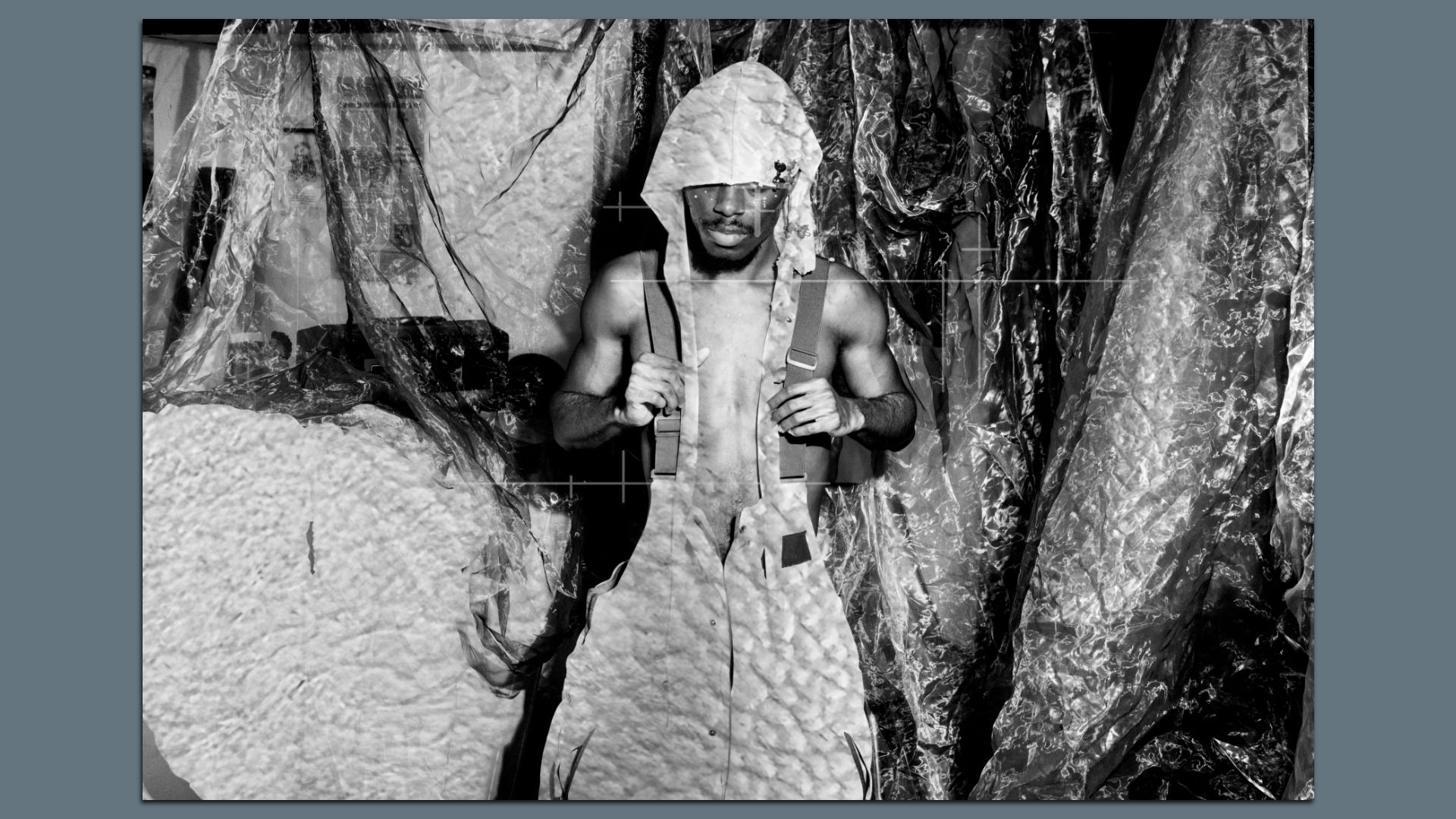Black-and-white artistic image of a man wearing a hood and suspenders. Standing in front of various fabrics with unique textures. 