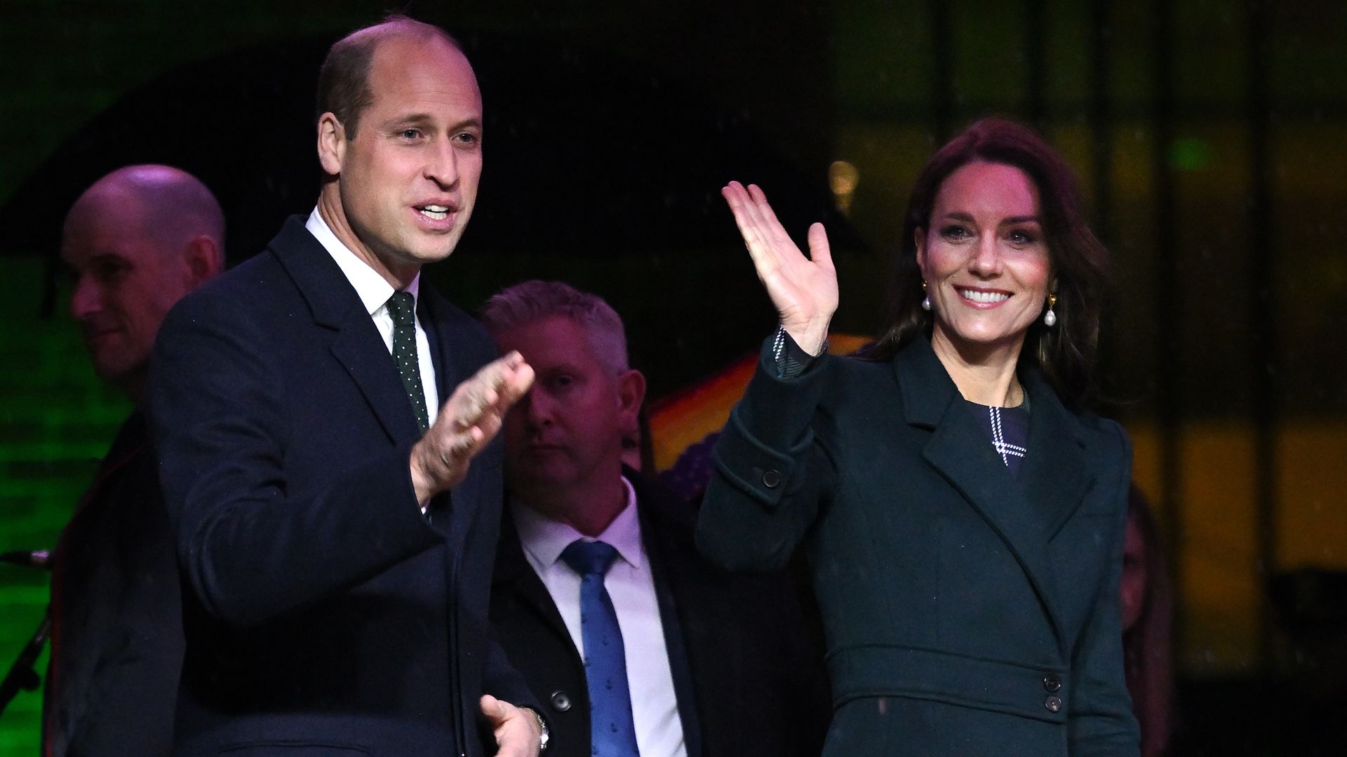 Prince William, left, and Princess Kate, right, wave as they stand by a podium with a green button. They pressed it and turned City Hall's lights green.
