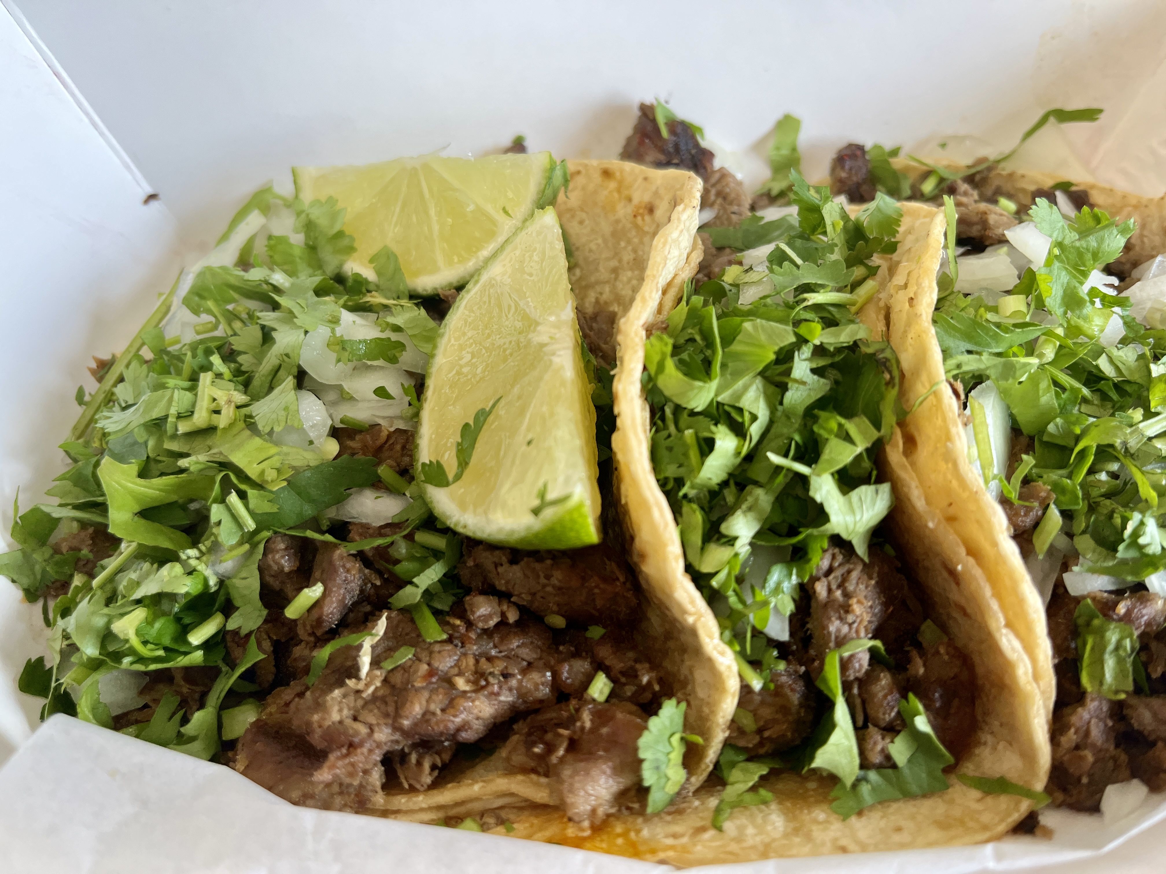 Three tacos filled with grilled meat, topped with chopped white onions and fresh green cilantro, served with lime wedges in a white container.