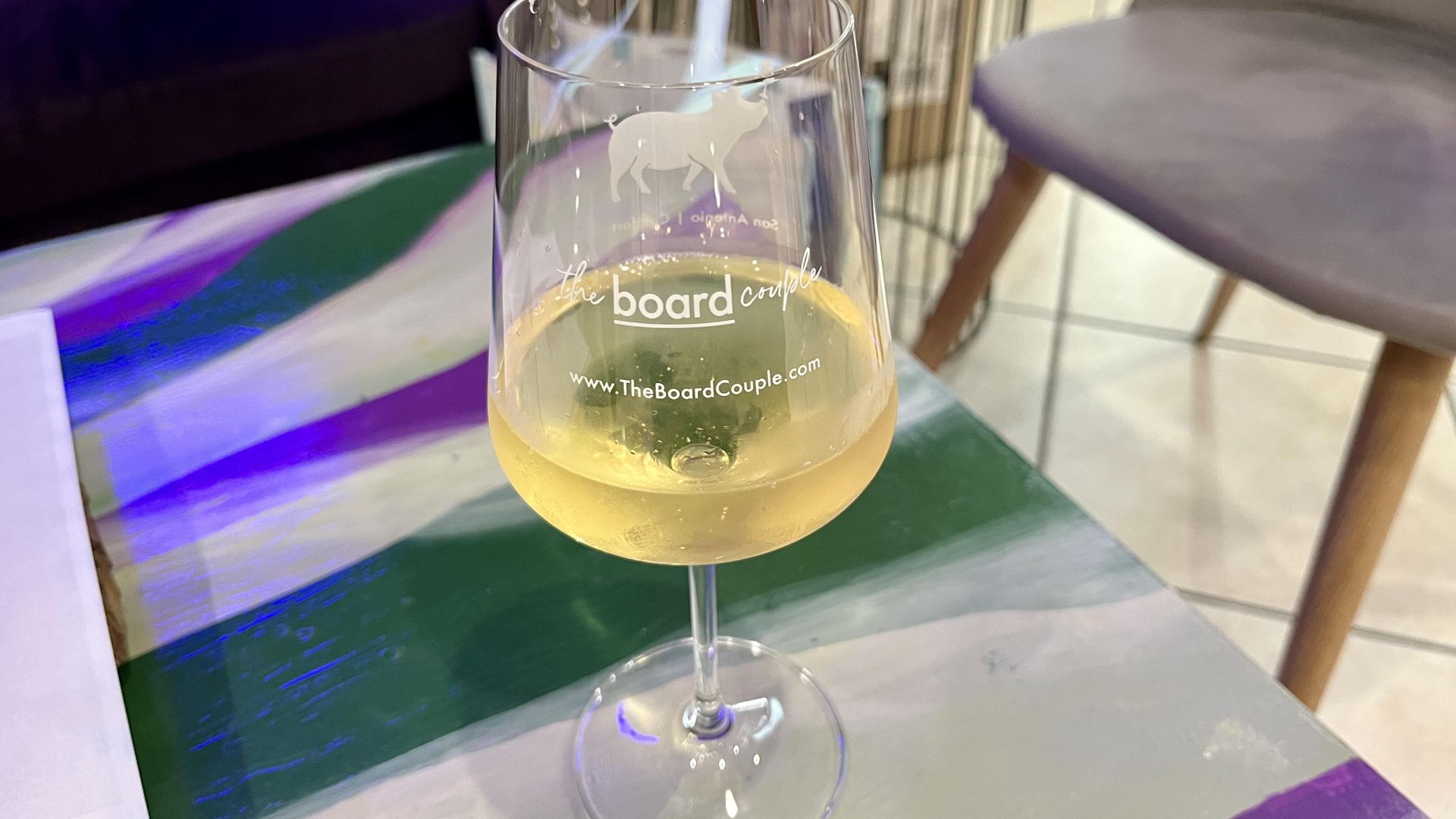 Wine glass with a pale yellow drink on a colorful striped table. The glass shows a white horse logo and the words "the board couple" with the site "www.TheBoardCouple.com".