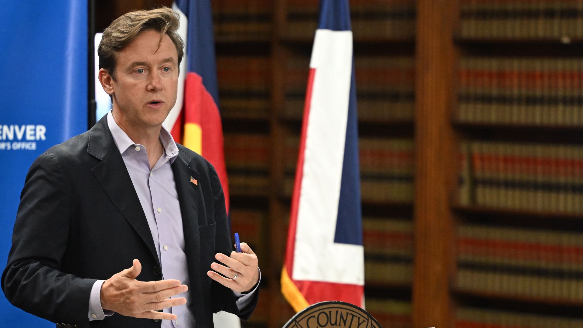 A photo of Mayor Mike Johnston speaking with his hands up at a podium with a state and city flag behind him.