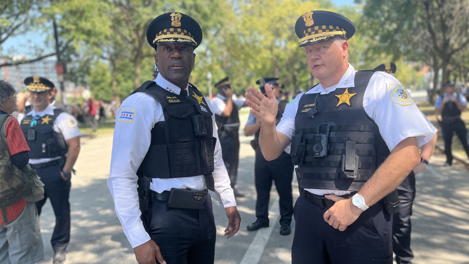Two police officers in white shirts and black tactical vests, wearing peaked caps with gold insignia, stand outdoors on a sunny day; trees and other officers in background.