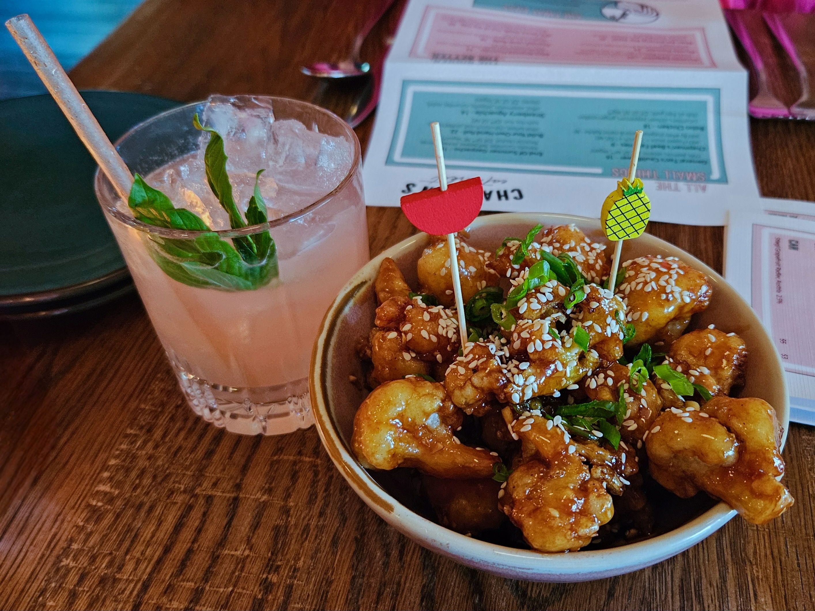 A pink margarita garnished with a leaf, next to a bowl of General Tso's cauliflower with two decorative toothpicks in it 