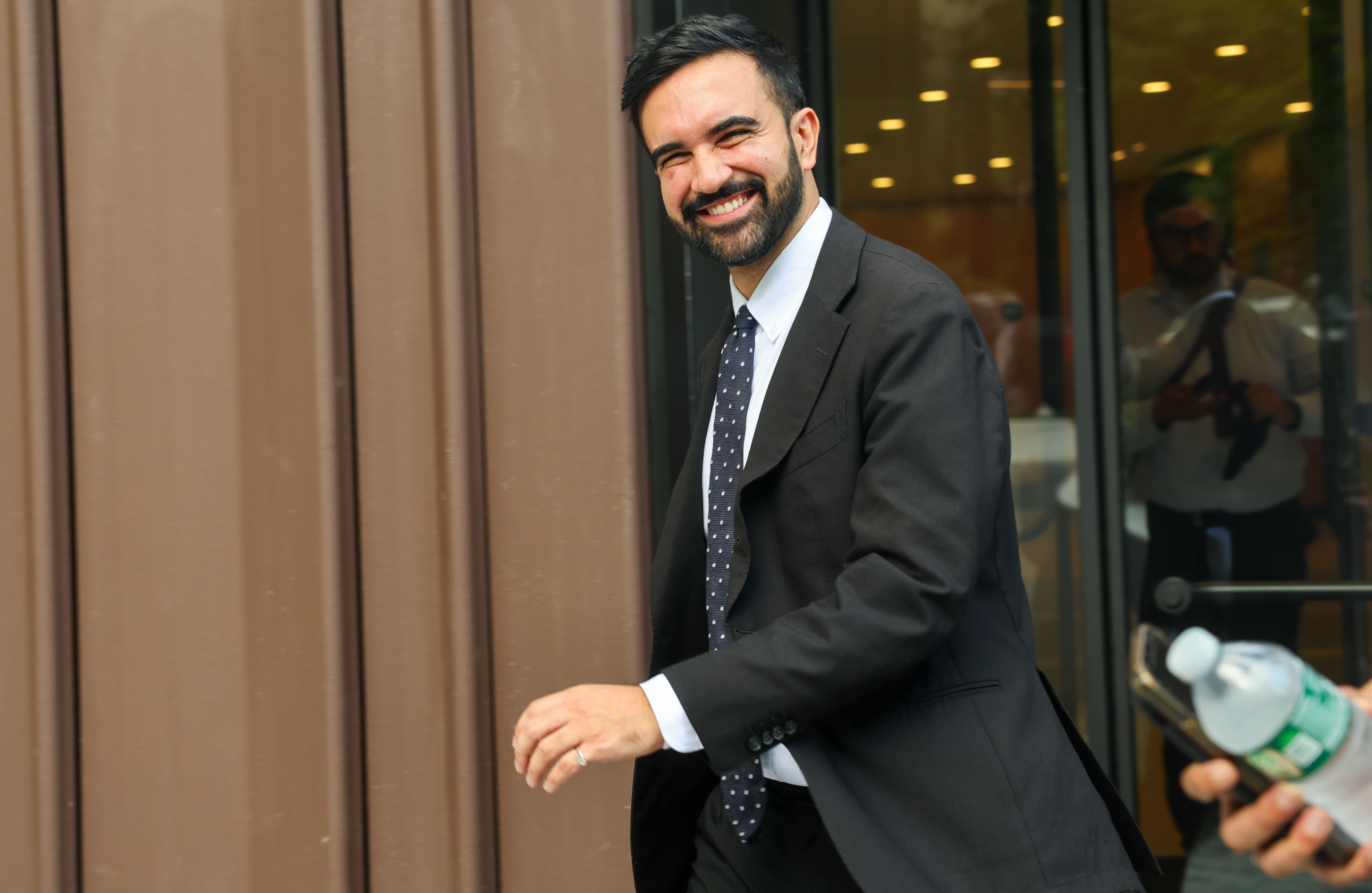 Smiling man with dark hair and beard wearing a black suit with a white shirt and navy blue polka dot tie, walking past brown wall and glass door, holding something in his left hand.