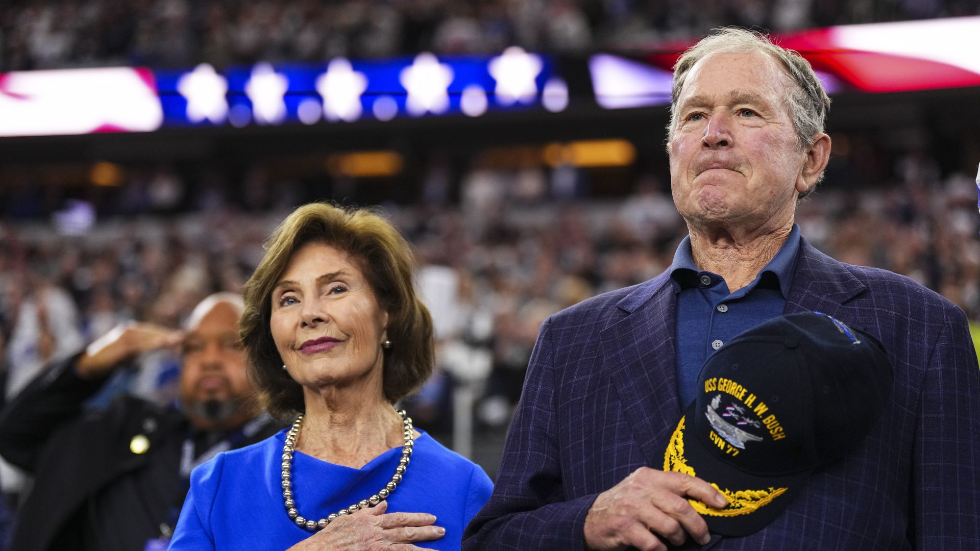 Former President George W. Bush and former First Lady Laura Bush stand with their hands over their hearts during a football game.