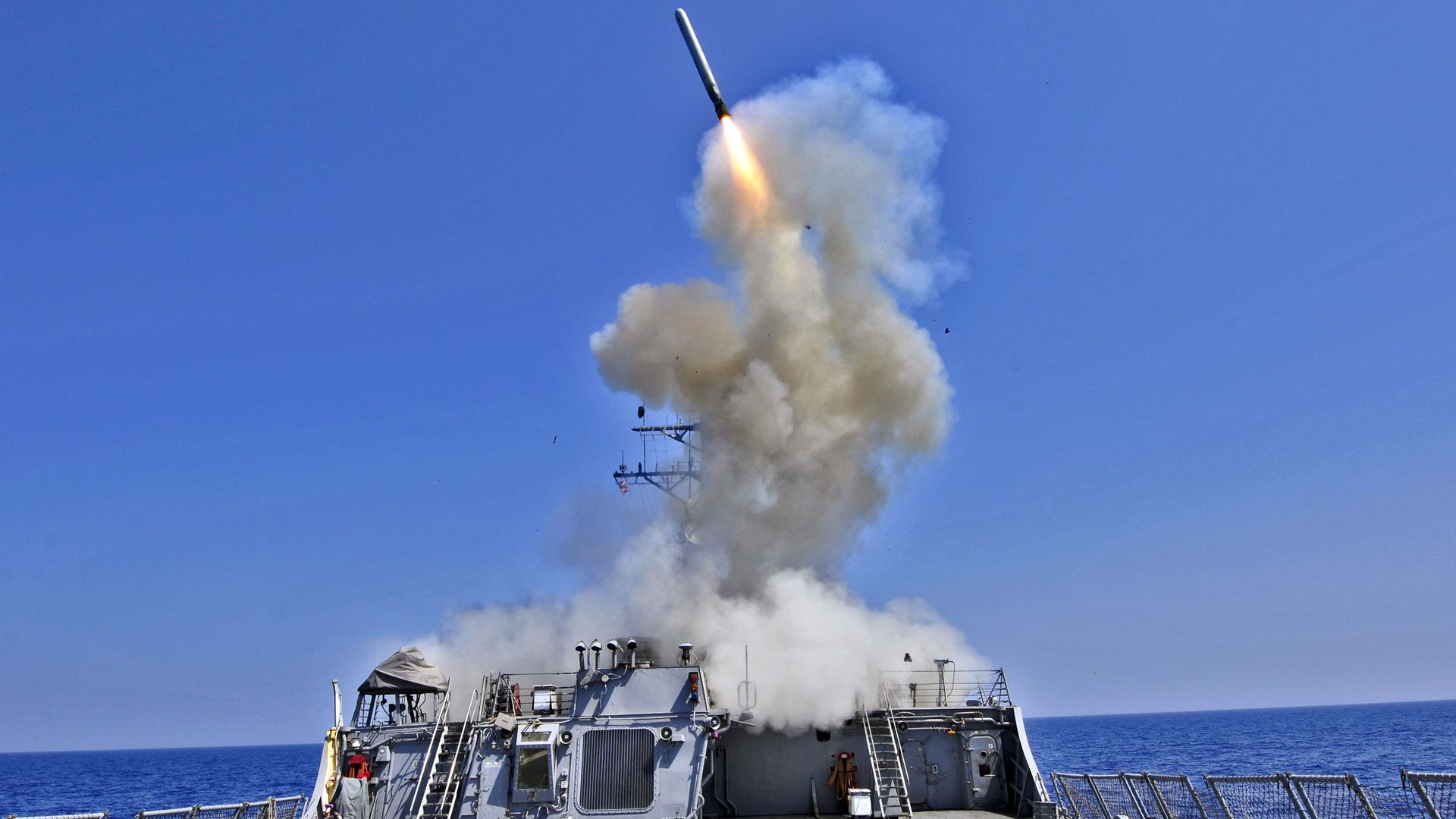 Missile launching from the deck of a gray naval ship amid blue sky and sea, with a large plume of white smoke rising from the launch point.