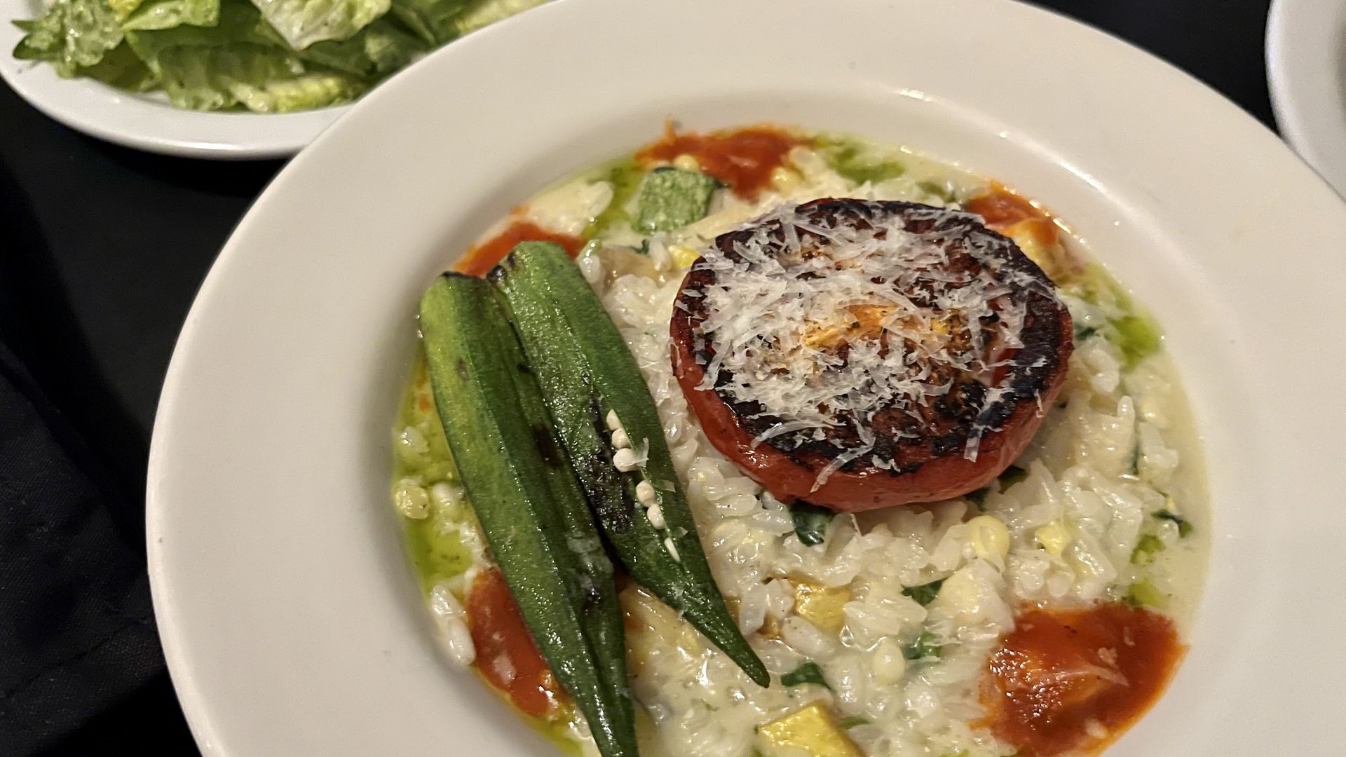 A white, shallow bowl holds creamy risotto topped with a charred tomato and grated cheese and two roasted okra pods. A side salad with green lettuce and drinks are seen in the background. This is a locally sourced summer dish from Outlaw Kitchens in recent years.