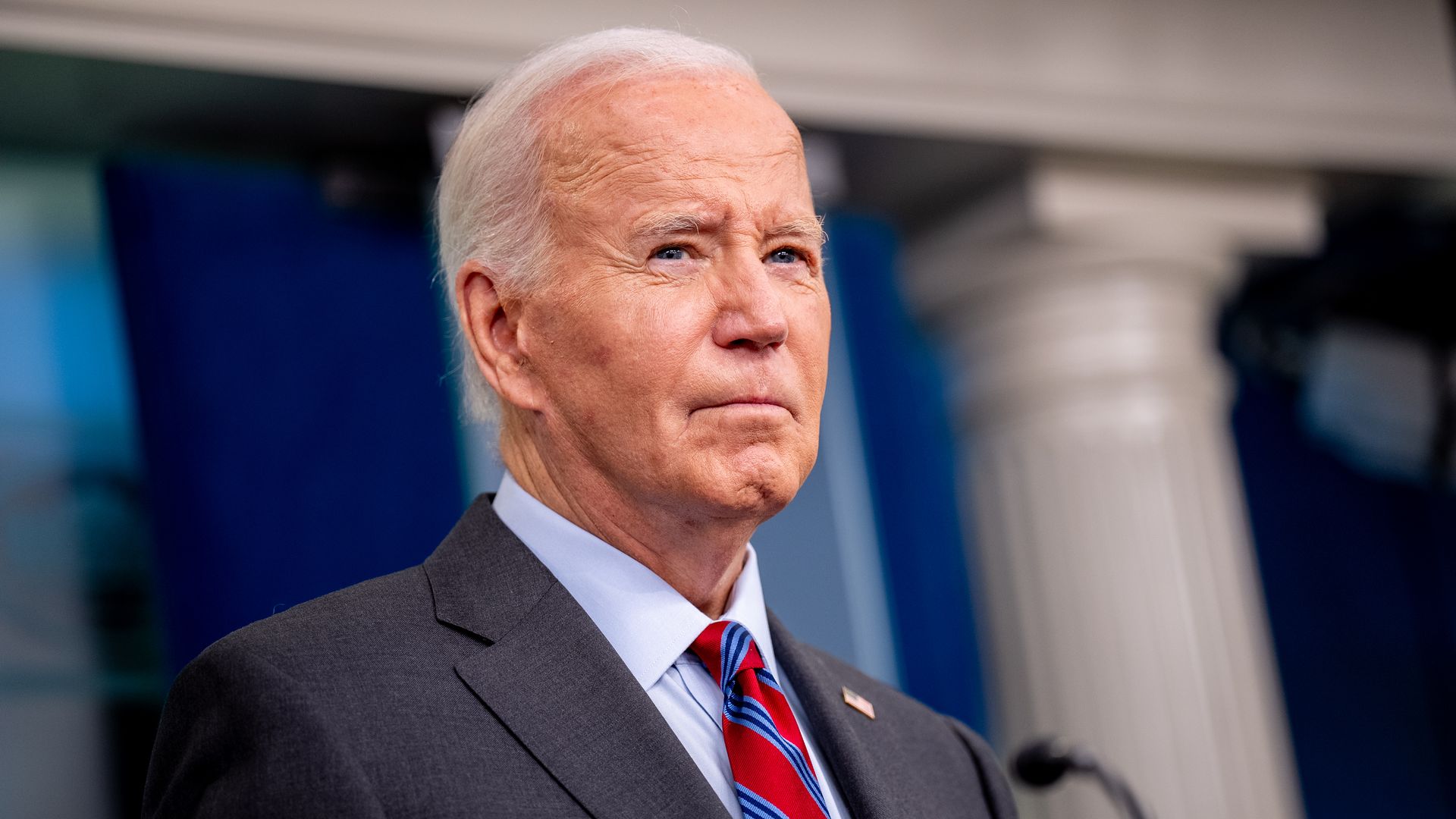 President Biden wearing a gray suit, white shirt and red and blue striped tie standing in the White House press briefing room.