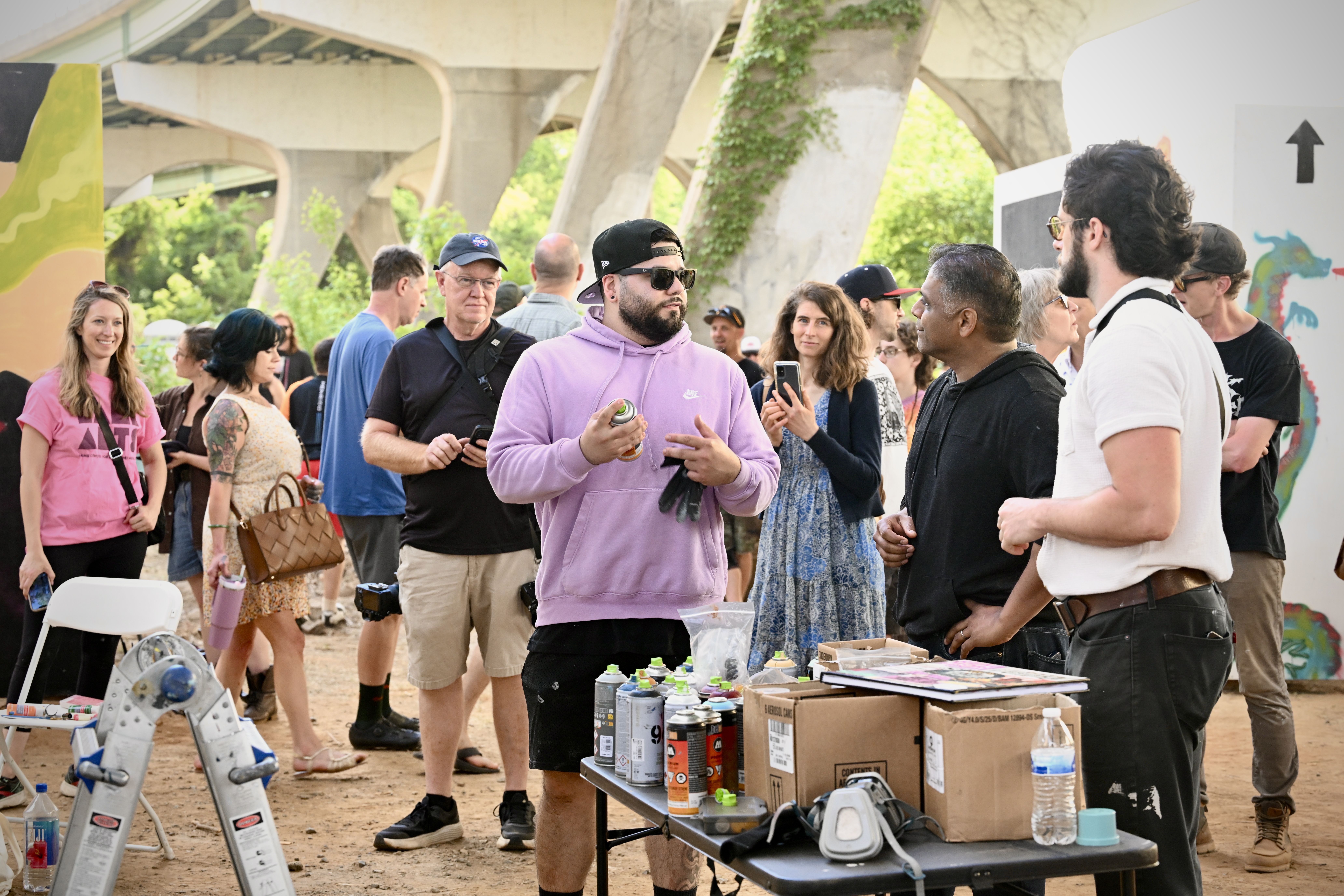Group of people at an outdoor event beneath a concrete bridge. In the foreground, a man in a lavender hoodie and cap holds a spray can; a table shows spray cans, boxes, and water bottles.