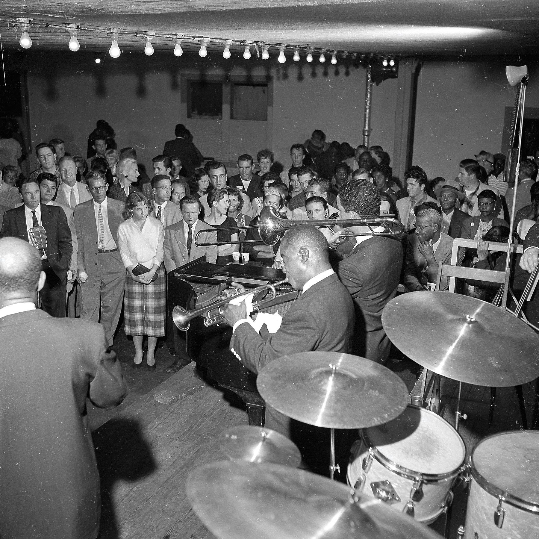 Black-and-white photo of a crowded 1950s-style party; a live brass band plays in front of a formally dressed audience under hanging string lights.