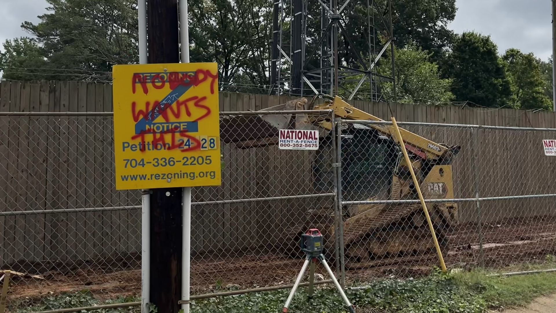 Construction site with yellow CAT bulldozer behind a chain-link fence, a wooden fence and trees in background, and a yellow rezoning notice sign vandalized with red graffiti reading "Nobody Wants This."