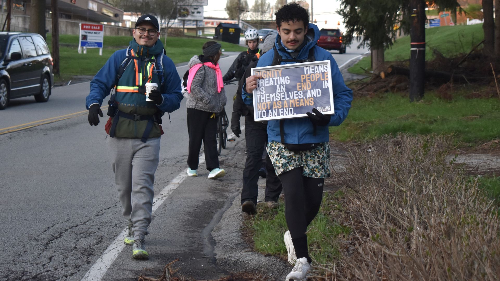 Two young protesters walk along a street, one in a blue jacket with a reflective vest, smiling with a cup, the other holding a sign about dignity; others and cars in the background.