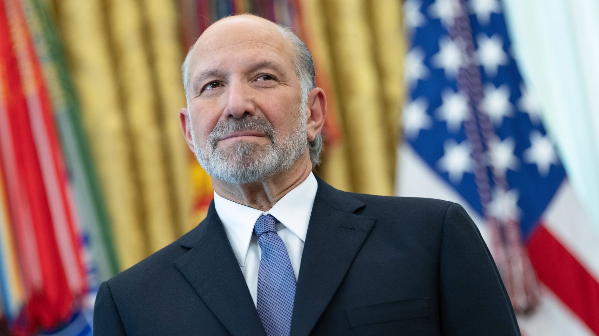 A man with a gray beard in a dark suit and blue tie stands in a formal setting, with colorful banners and part of the American flag in the background.