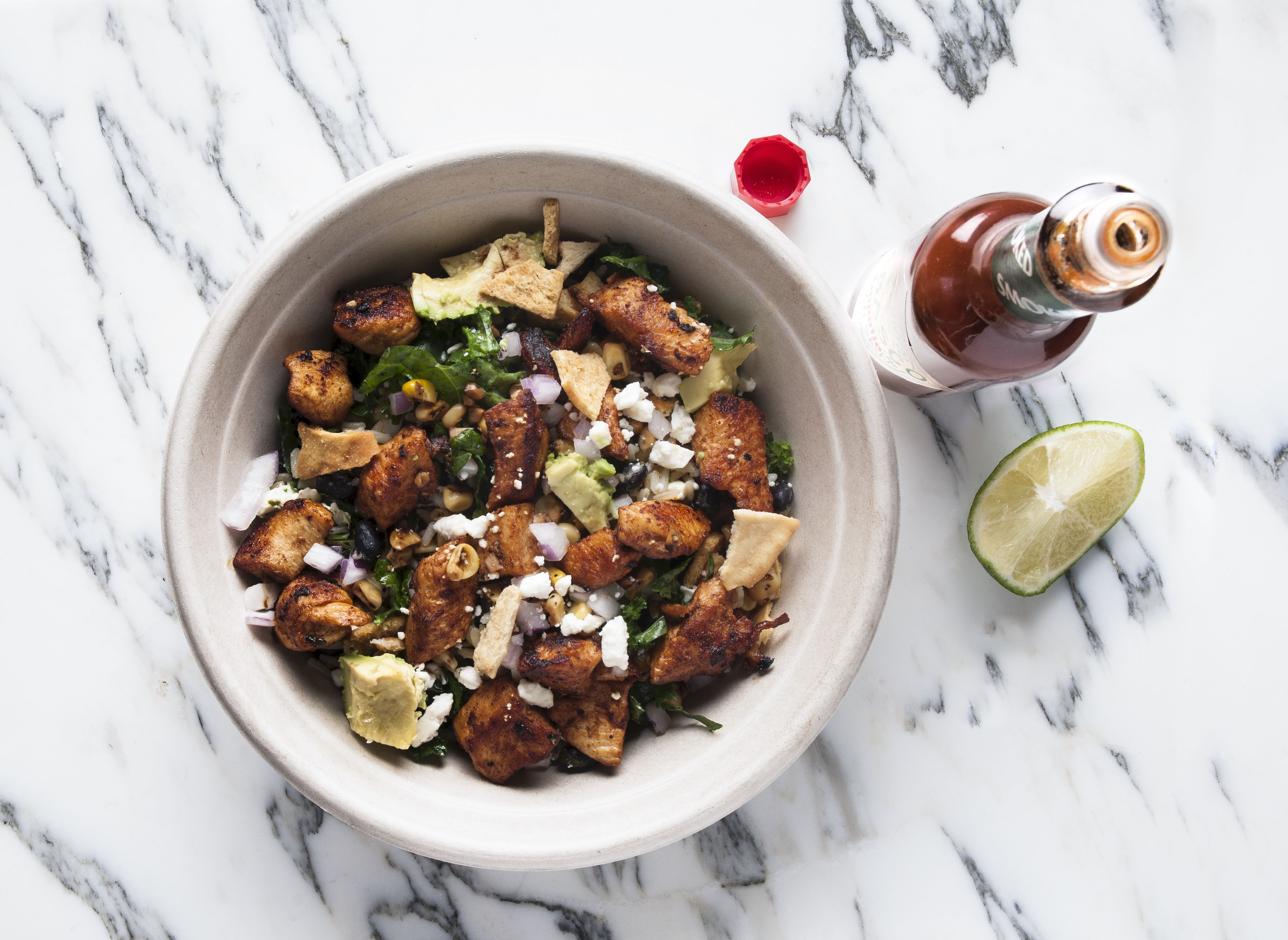 Top-down shot of a beige bowl with a mixed salad: grilled chicken pieces, feta, greens, corn, olives, onions, and tortilla chips; a lime wedge and hot sauce bottle on a white marble surface.