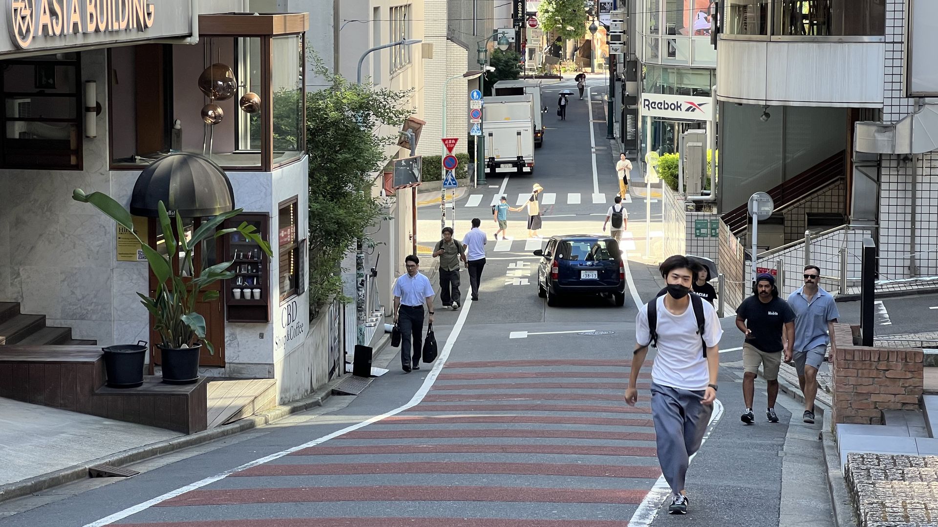 Busy urban street scene with pedestrians walking, cars passing, a building with "ASIA BUILDING" sign, and shops including Reebok, under bright daylight.