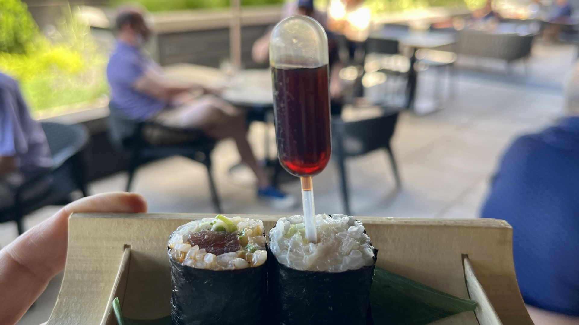 Close-up of two sushi rolls wrapped in nori, held by a hand. A small glass of red drink on a stick sits behind, with a blurred outdoor cafe and people in the background.