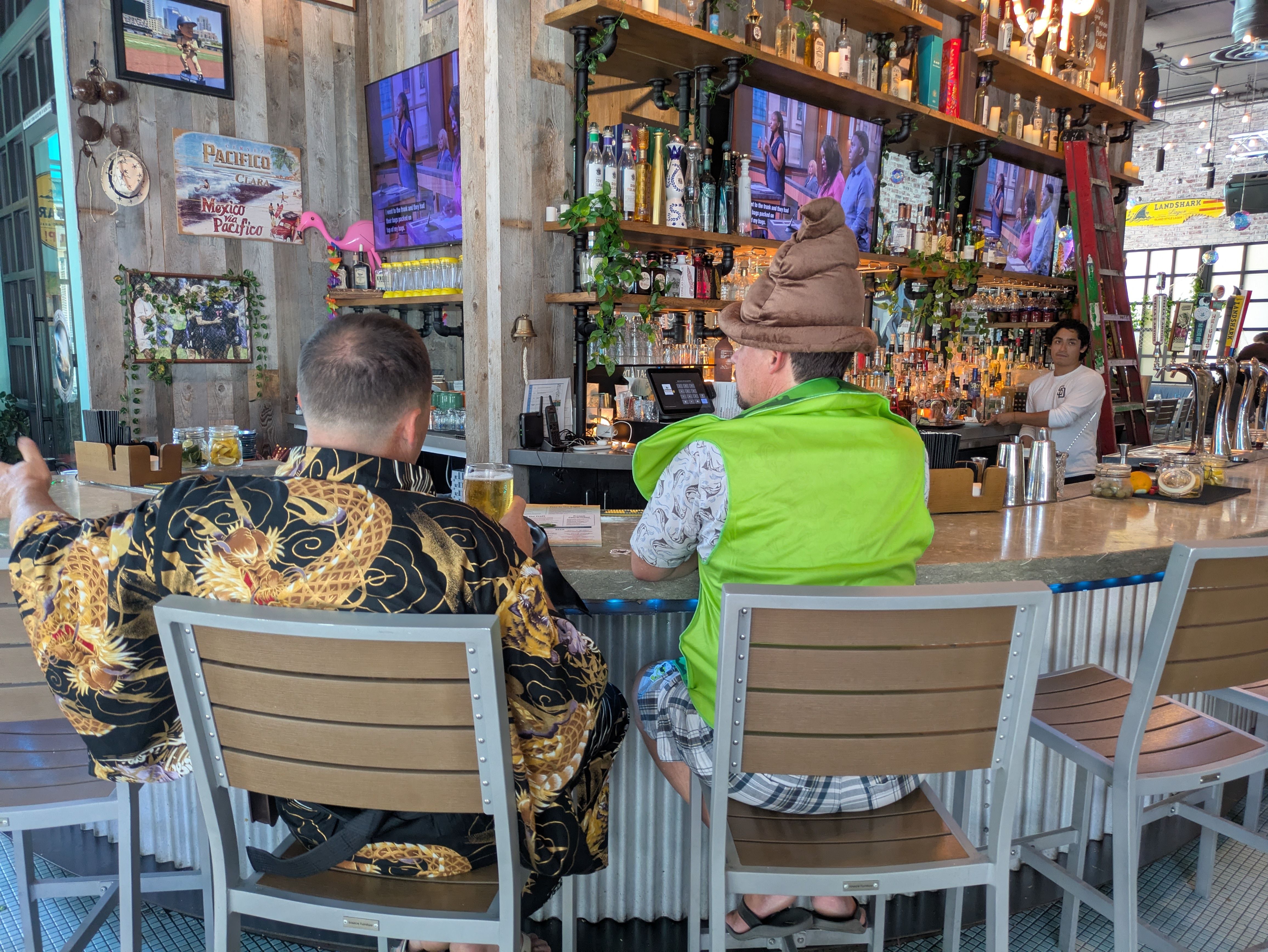 Bar interior with wood-paneled walls, liquor shelves, and several TVs. Patrons in tropical shirts sit at a curved counter; a man in a brown hat faces the bar as a red ladder sits nearby.