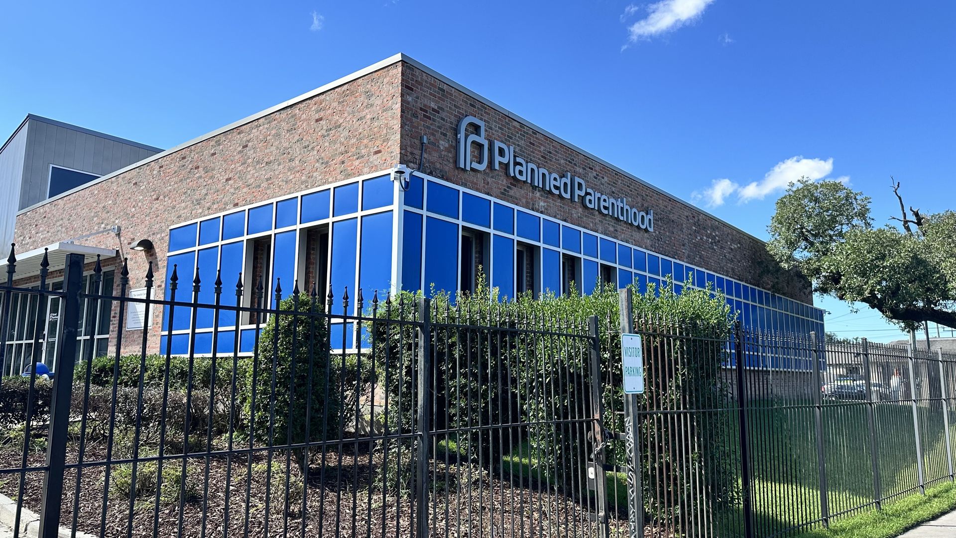 Exterior of a Planned Parenthood building with brick walls, blue glass windows, a black iron fence in front, shrubbery, and a clear blue sky.