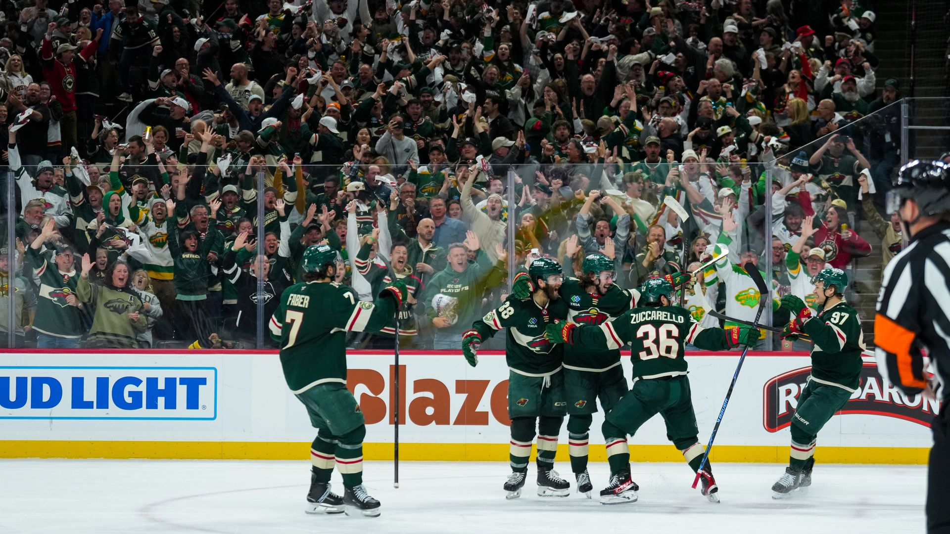 Large hockey crowd in a packed arena celebrates; players in green skate near the boards, raising arms and sticks, as fans cheer behind the glass and a referee stands in the foreground.