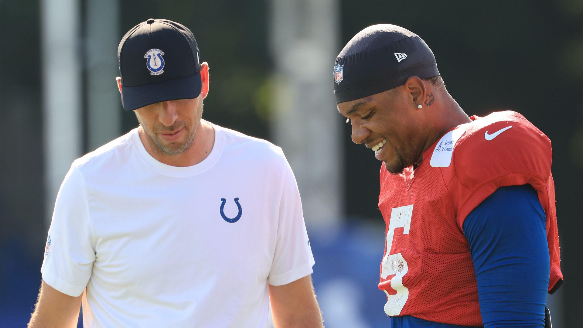 Two men outdoors at a football practice; one in a white Colts T-shirt and black cap, the other in a red practice jersey with number 15 and a black NFL cap, both smiling and looking down.