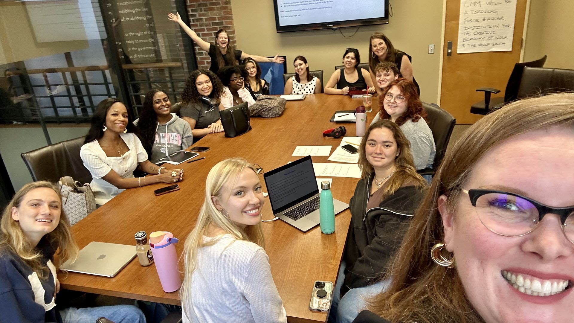 Group of diverse young adults smiling around a conference table with laptops and notebooks in a meeting room, one person takes a close selfie. A TV and a handwritten poster are on the walls.