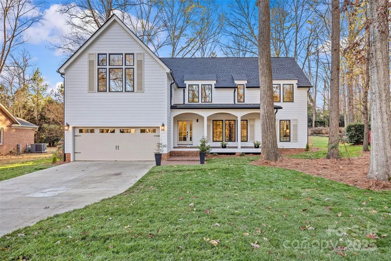 Two-story white house with gray roof, large garage, and front porch with potted plants. Surrounded by green lawn and tall trees under a blue sky.