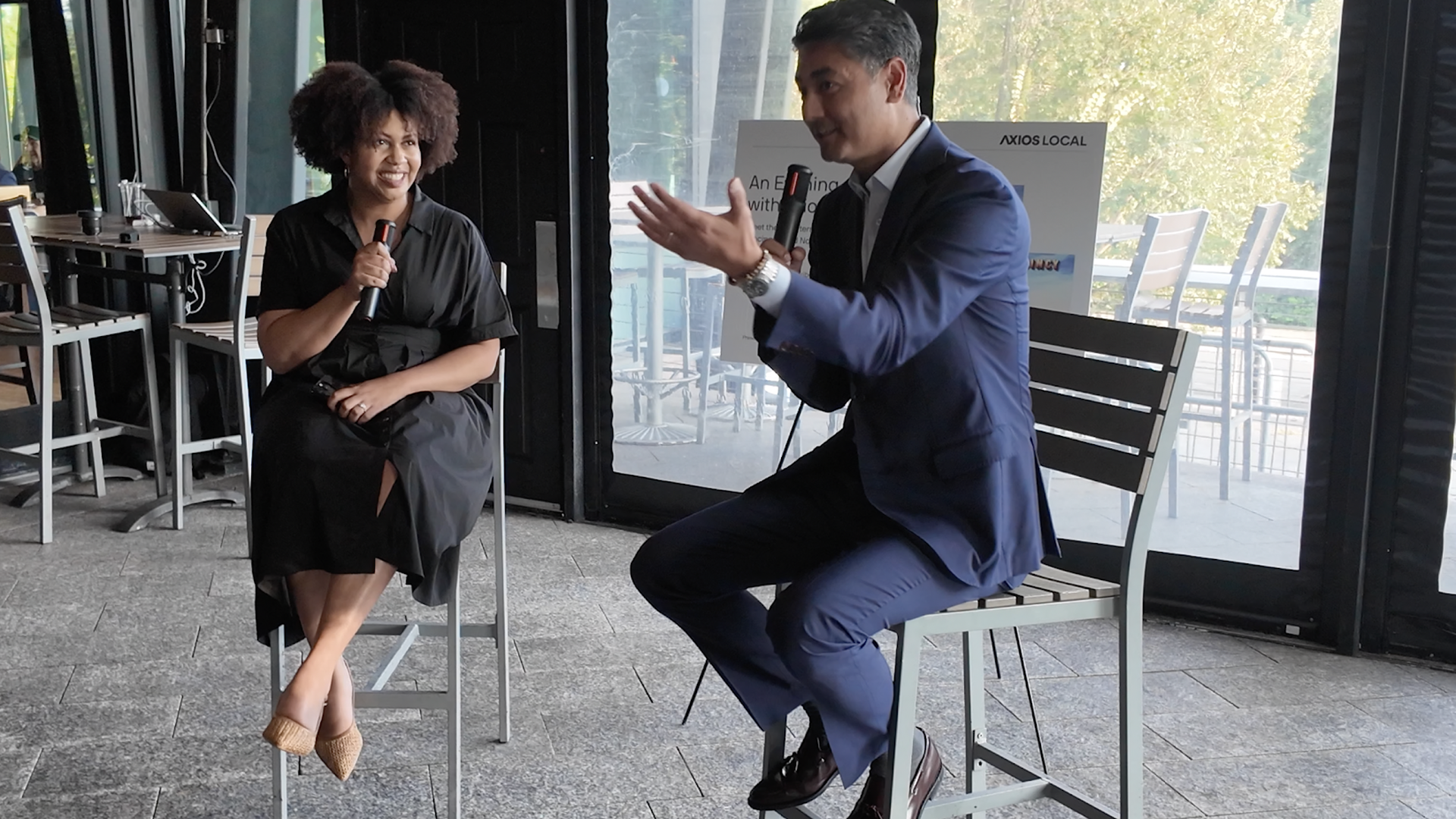 Mayor Aftab Pureval sits on a chair talking into a microphone while speaking to Axios Cincinnati reporter Kristen Swilley, also holding a microphone. They're in bar room with glass doors behind them.