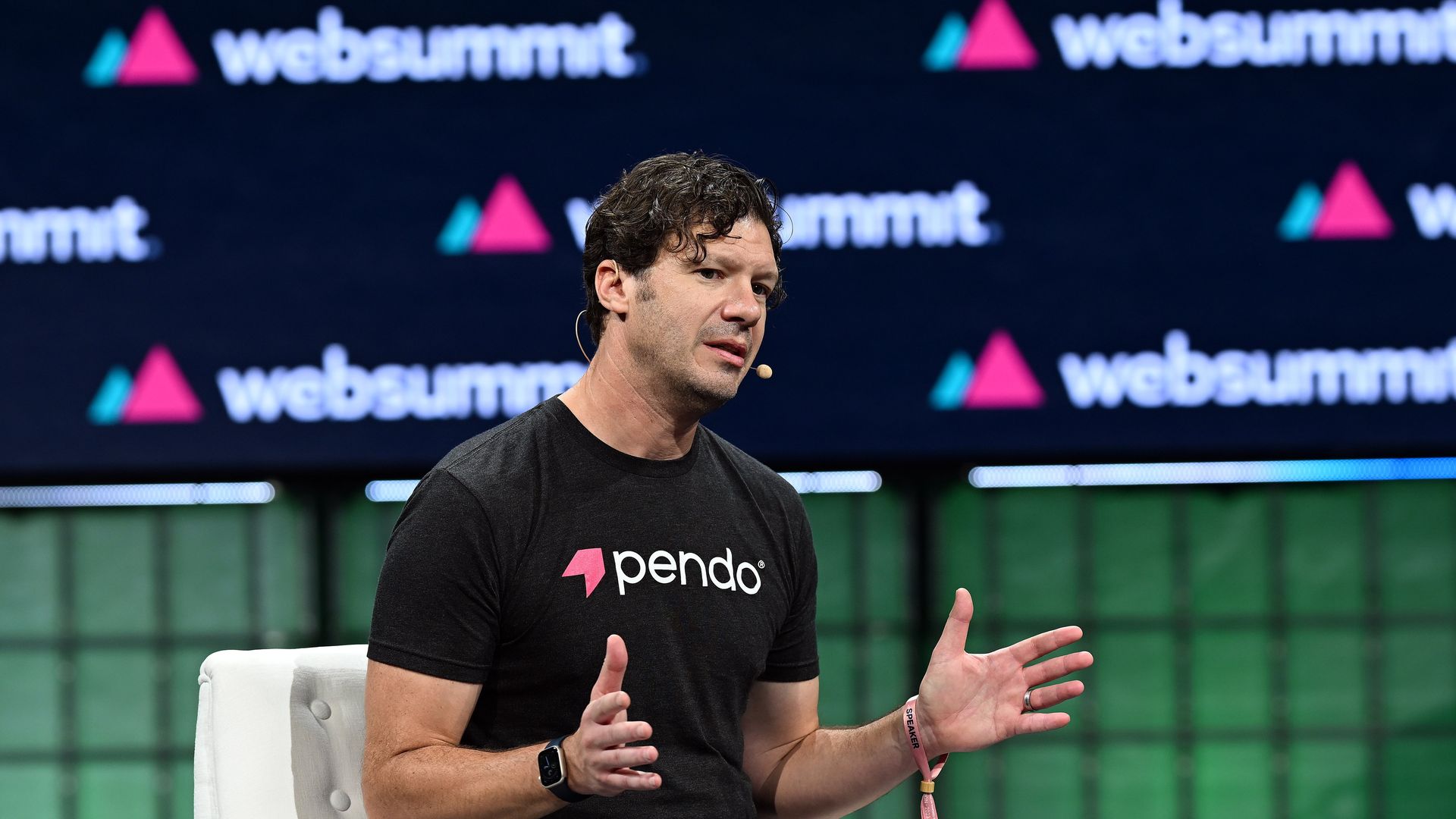 Man in a black pendo shirt sits on a white chair on stage, speaking with a headset mic and gesturing; dark backdrop shows "Web Summit" logos.