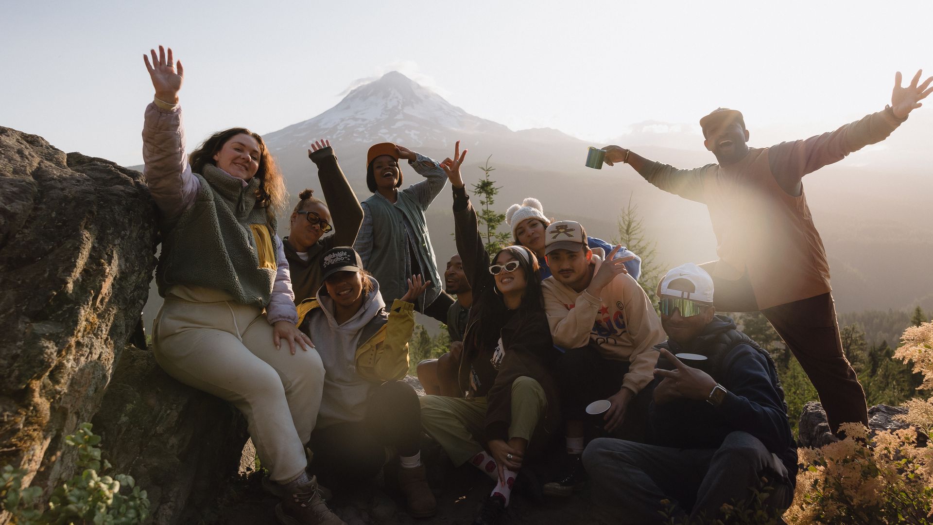 A group of people sit at the edge of a mountain with the snow-capped peak of another in the background.