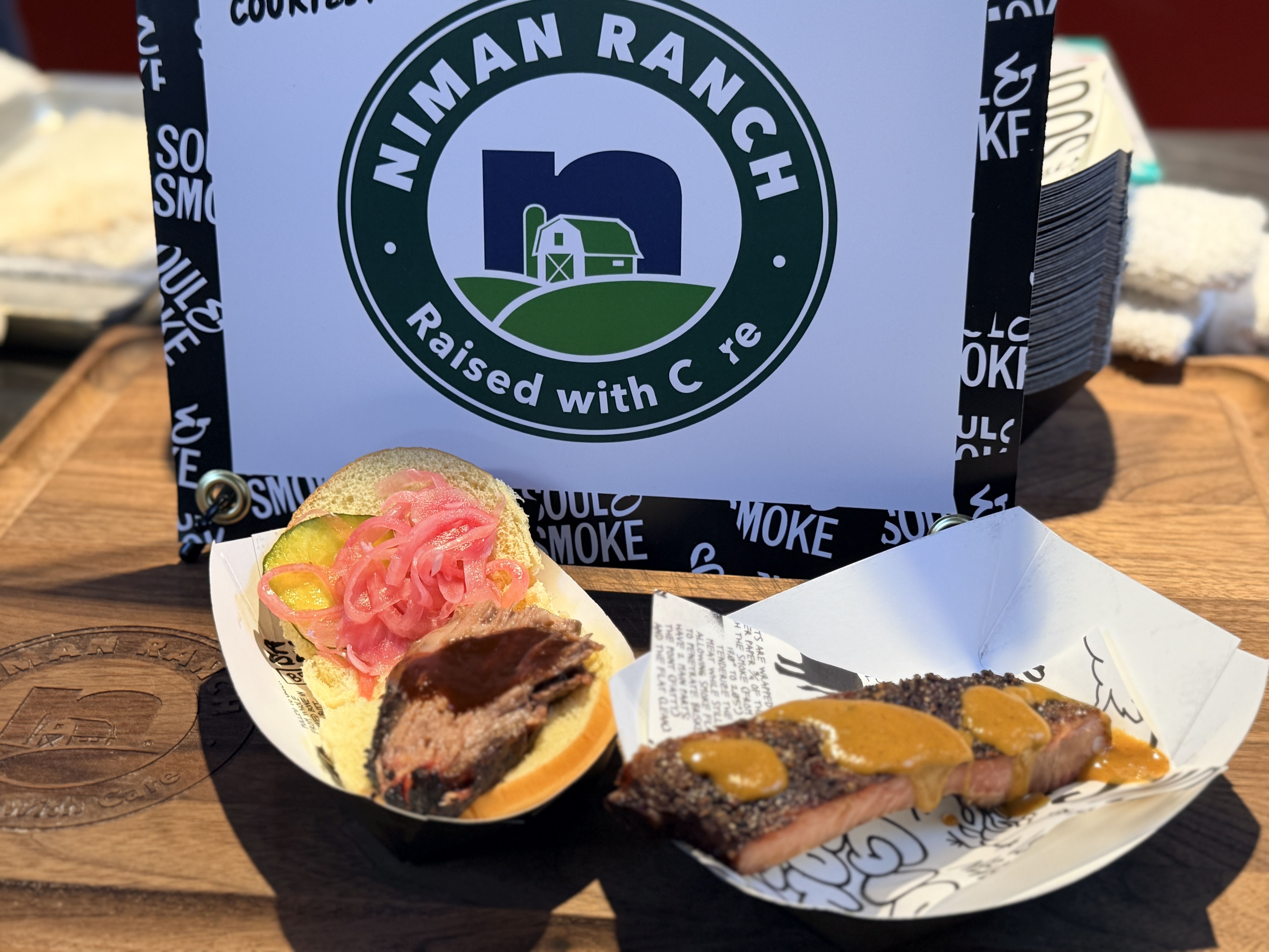 Outdoor scene with a white box featuring the green circular Niman Ranch logo (barn and silo). In front are pink onion-topped sliders on a wooden table.