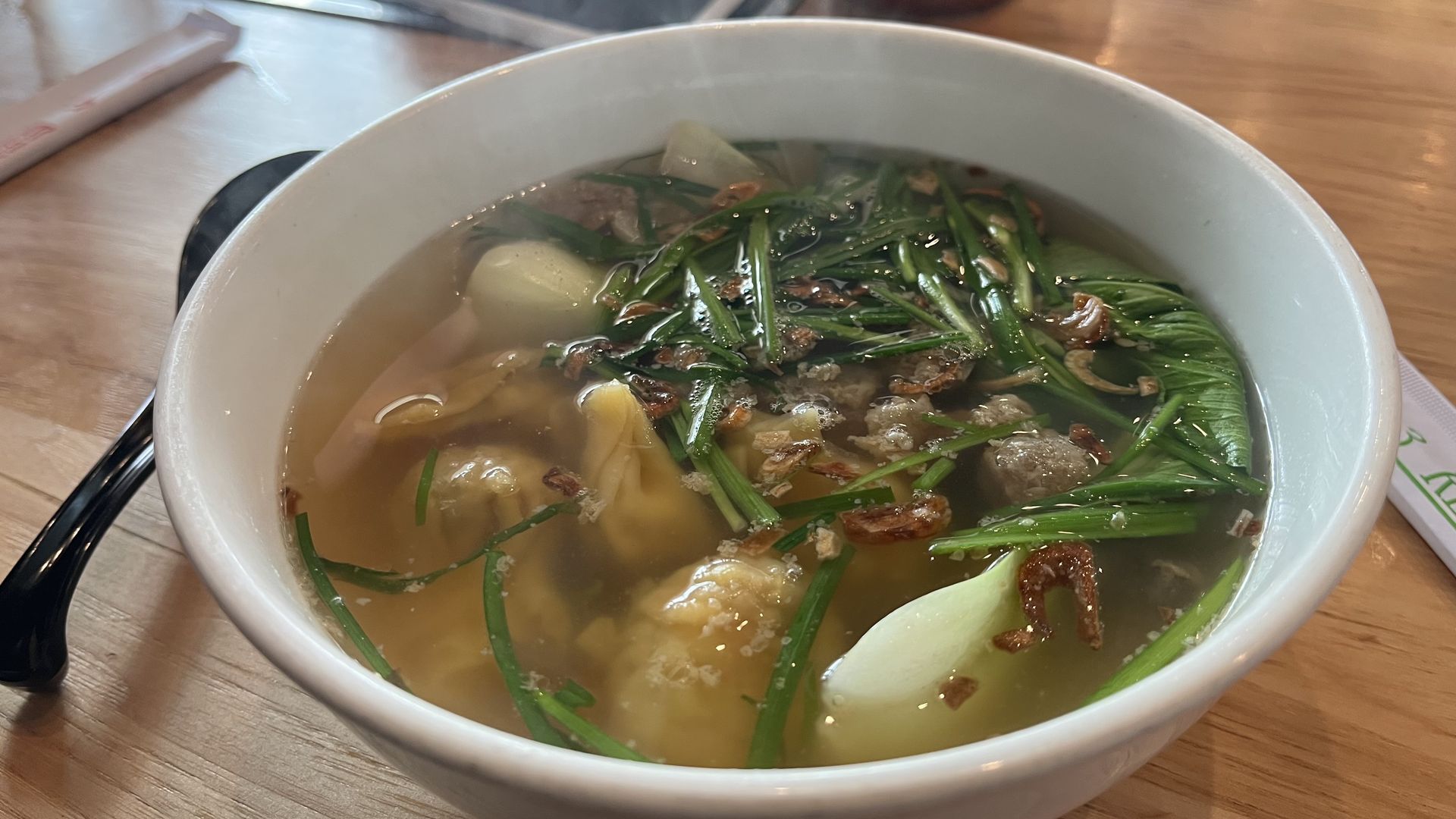 A white ceramic bowl of clear broth soup topped with leafy greens and fried shallots, with dumplings visible; black chopsticks and a white spoon sit on a wooden table.