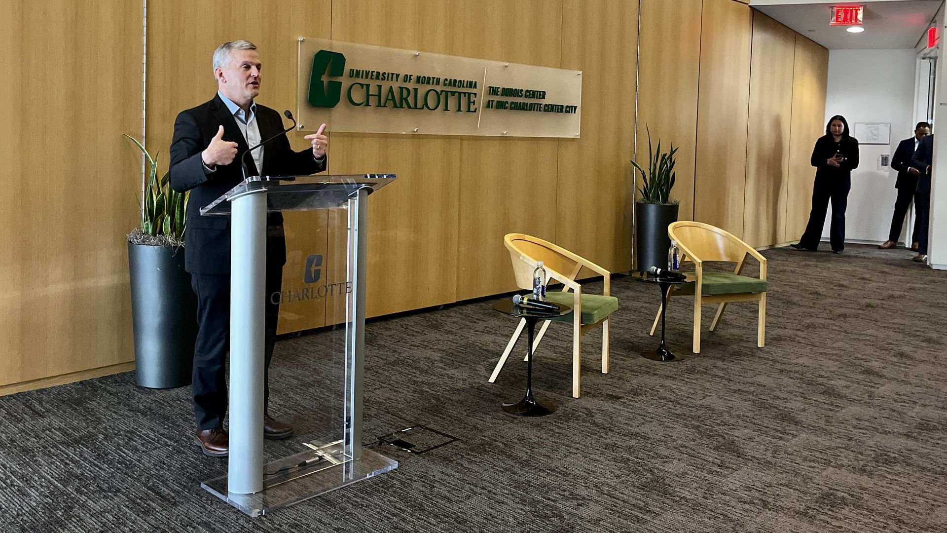 Speaker in a dark suit at a clear podium gives a talk in a wood-paneled UNC Charlotte conference room; two light-colored chairs, small tables with water bottles, and a few people near the back.