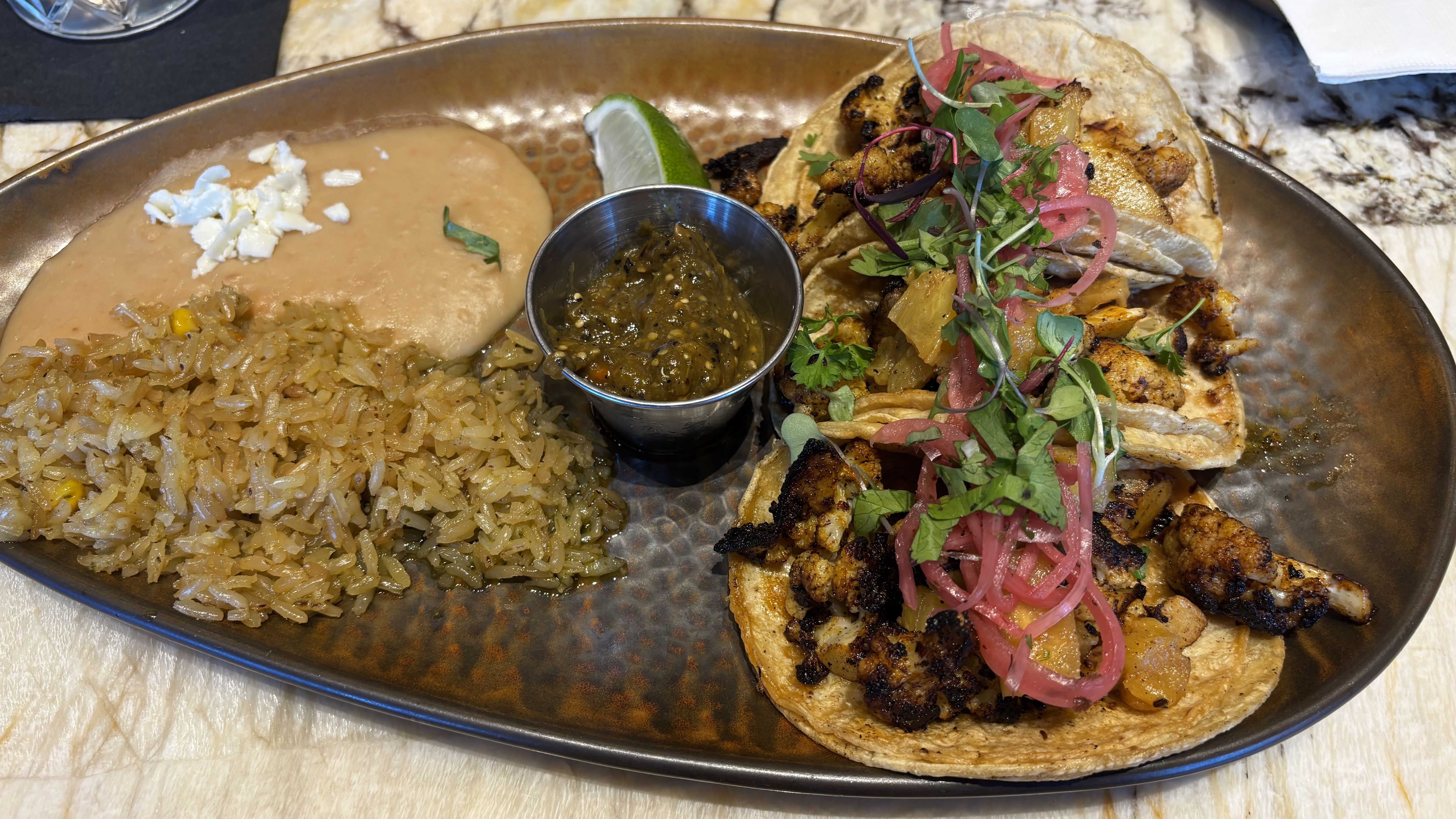 Plate with three grilled cauliflower tacos topped with pickled onions and herbs, served with seasoned rice, refried beans topped with cheese, a lime wedge, and green salsa in a metal cup.