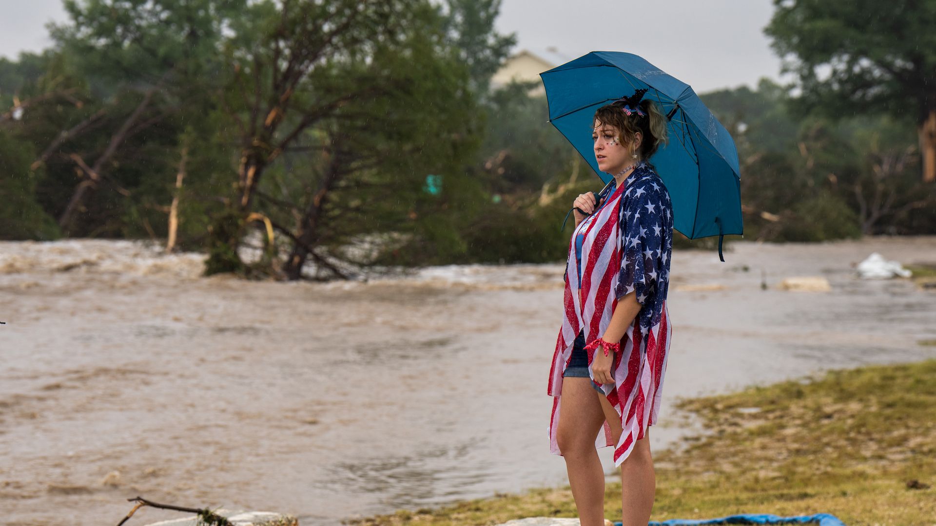 A woman with an america flag outfit draped over her holds an umbrella while standing next to floodwaters in Kerrville, TX