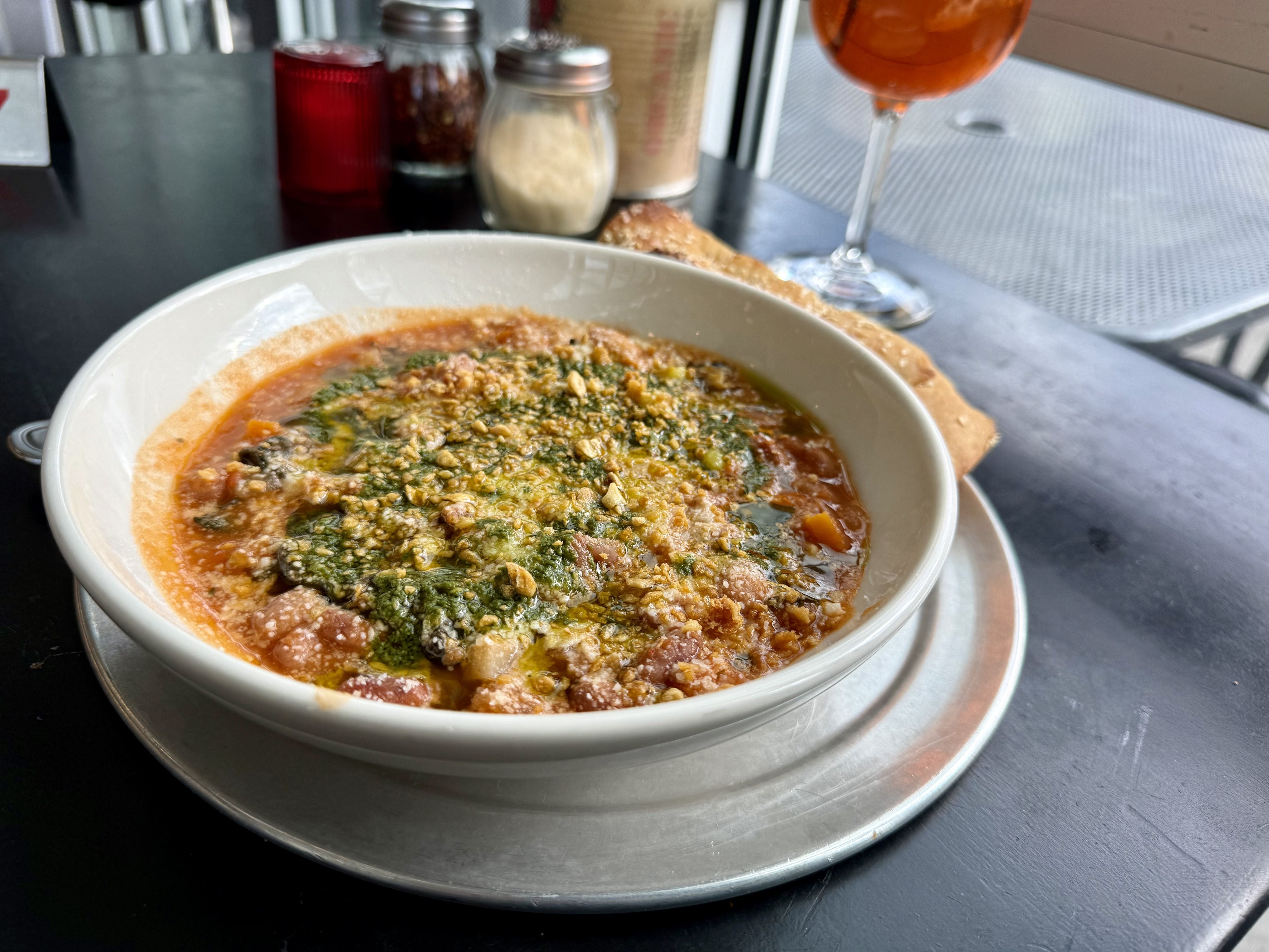 Bowl of tomato-based bean and vegetable soup topped with green herbs and grated cheese, served with bread on a silver plate on a black table next to a glass of amber drink.