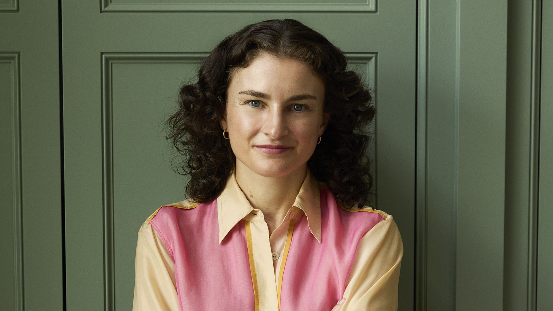A woman with curly dark hair stands with arms crossed, wearing a sheer pink and beige blouse and blue jeans, against a green paneled door with gold handles.