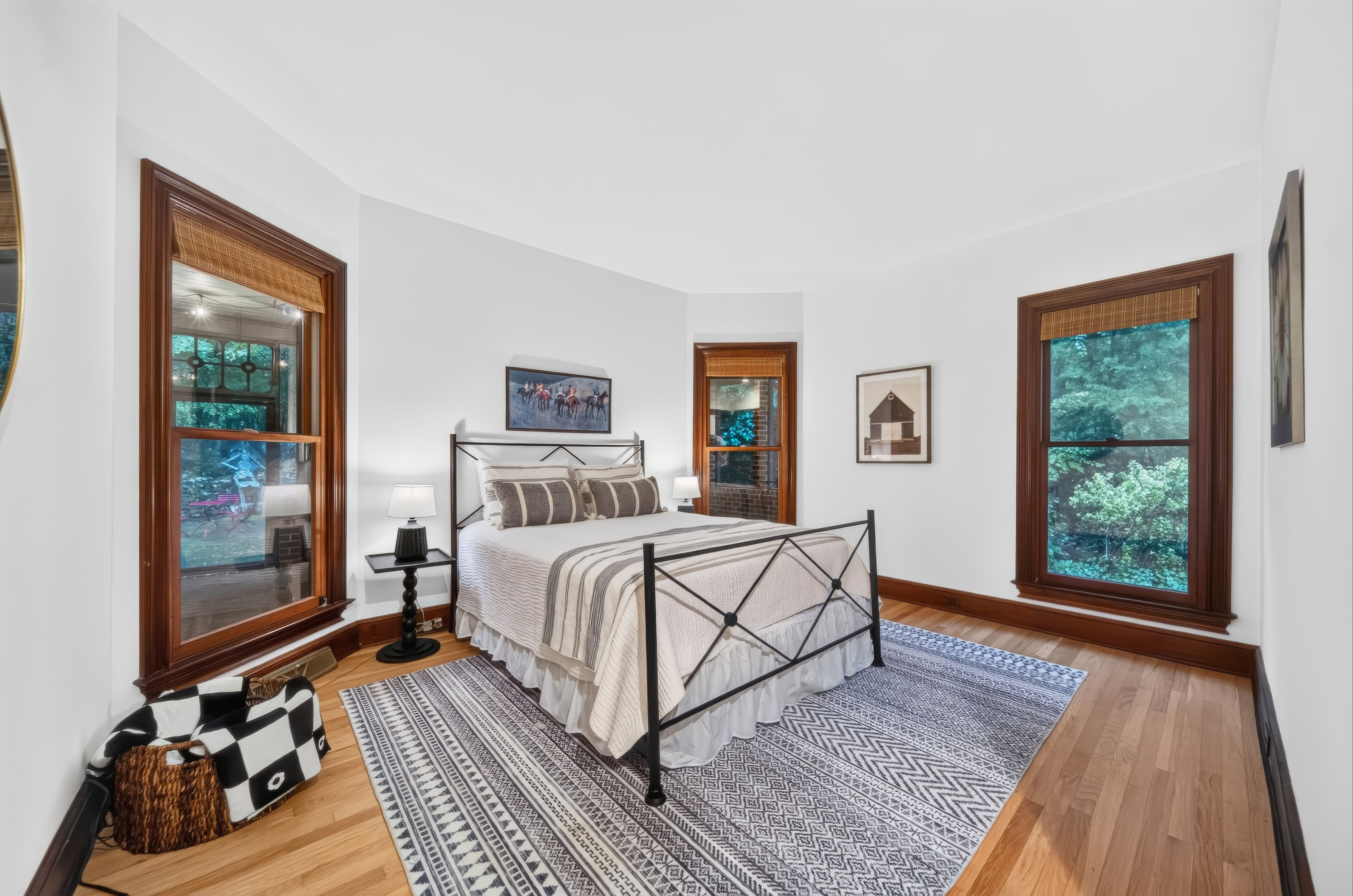 Bright bedroom with white walls, dark wood window trim, and a black metal bed frame with striped gray bedding. Two lamps flank the bed; a black-and-white rug and framed art complete the space.