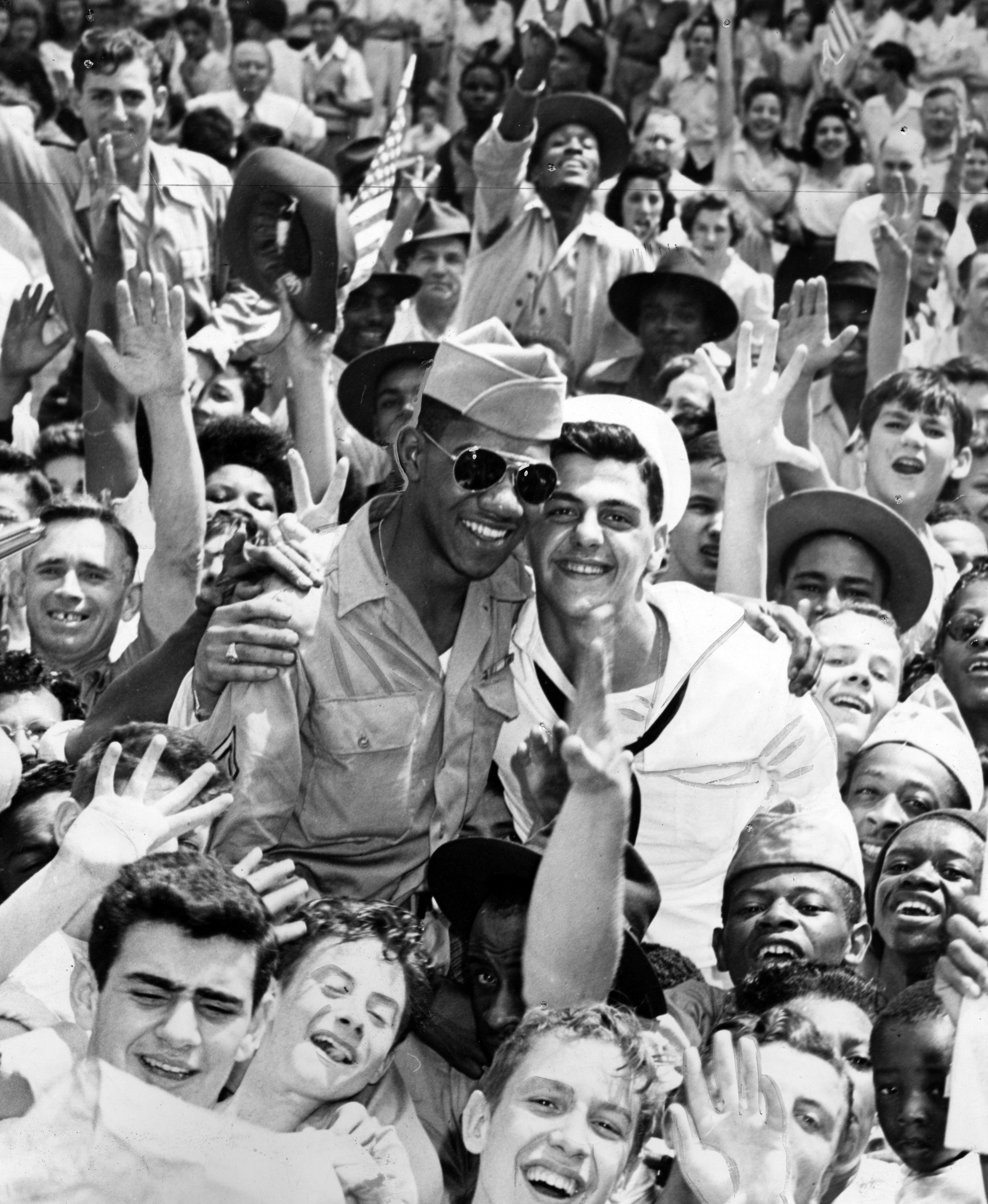 Black and white photo showing a joyful crowd of diverse military personnel and civilians, with two men in uniform smiling at the center, surrounded by raised hands and happy faces.