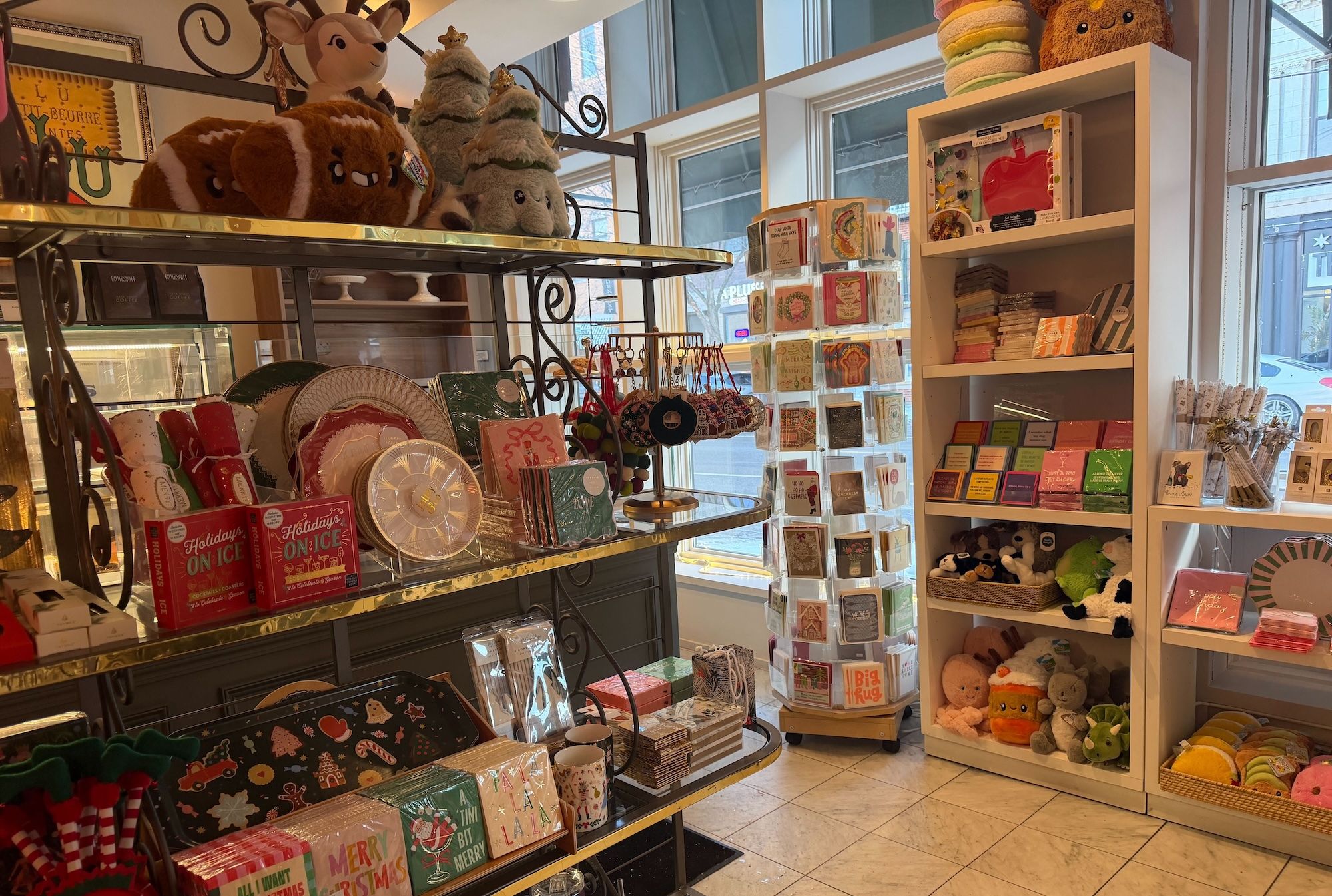 Inside Bittersweet, a gift shop displaying holiday-themed items including plush toys, decorative plates, cards, and colorful packaged goods on shelves and racks near large windows.